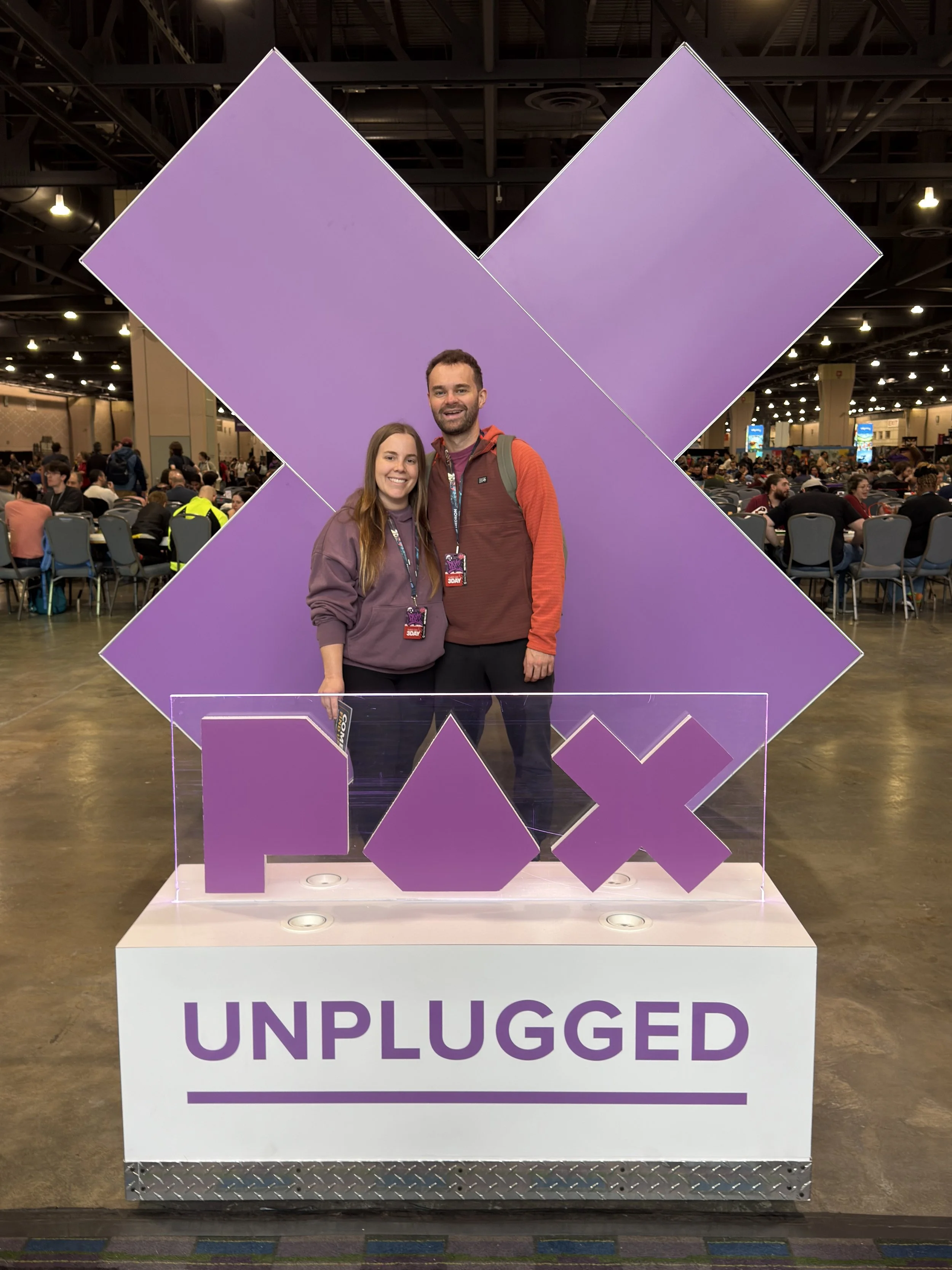 A smiling woman and man standing together at the PAX Unplugged event, behind a display with large purple icons and the word 'UNPLUGGED' in purple letters, with a crowded event space in the background.