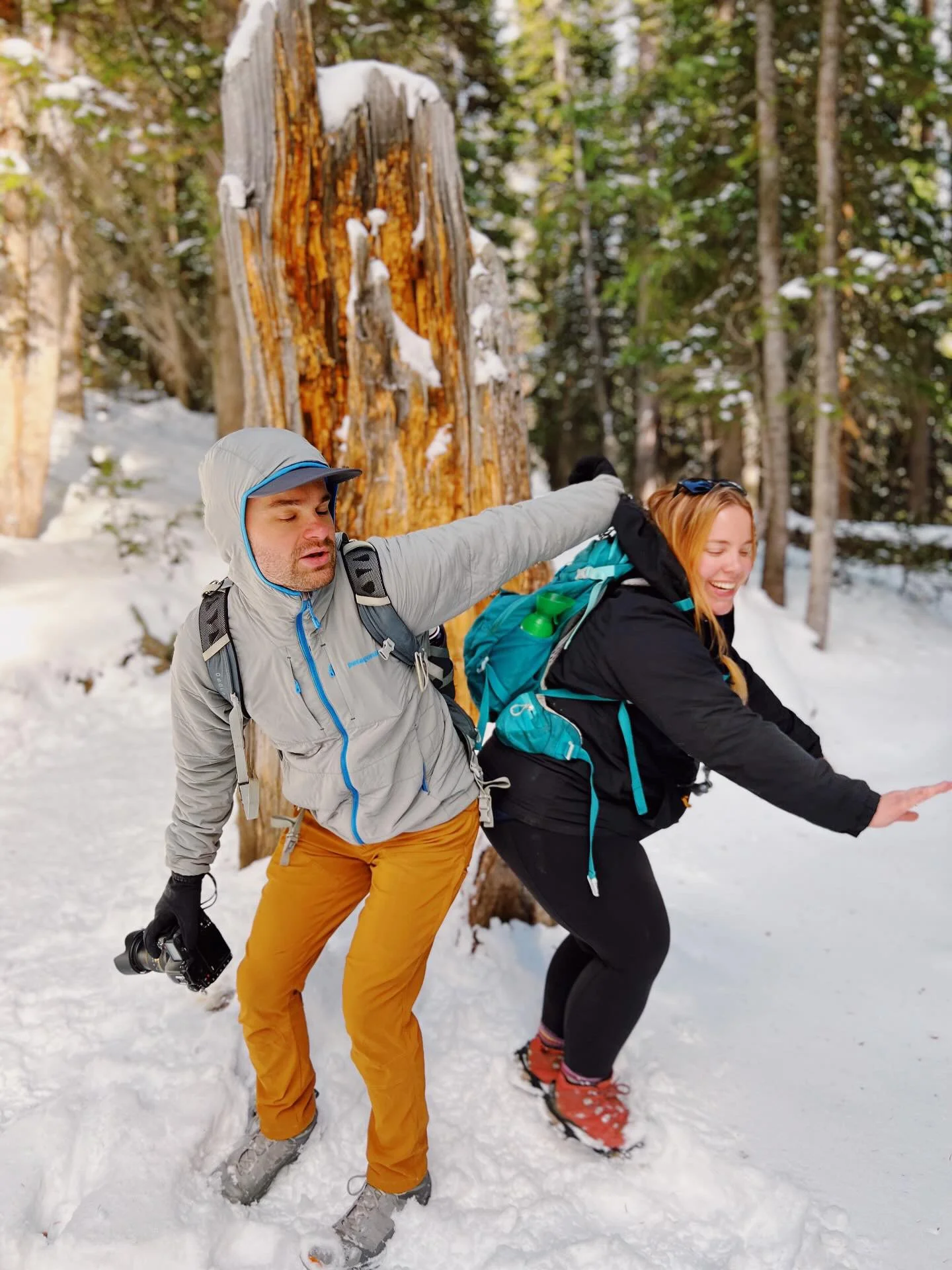 A man playfully grabbing a woman from behind in a snowy forest with trees and a large tree trunk in the background.