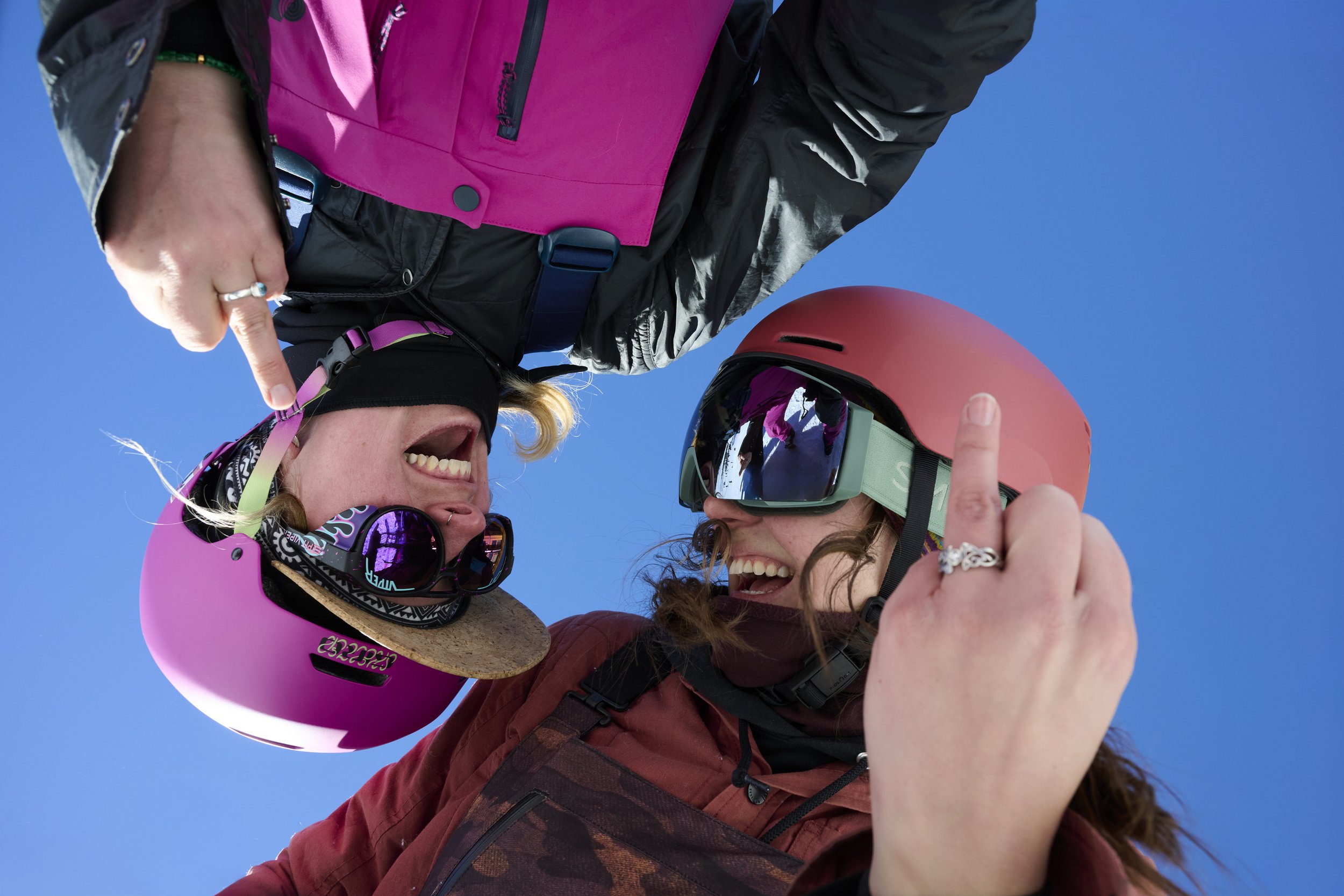 Two women with ski gear, smiling and laughing, wearing pink helmets and goggles, against a blue sky.