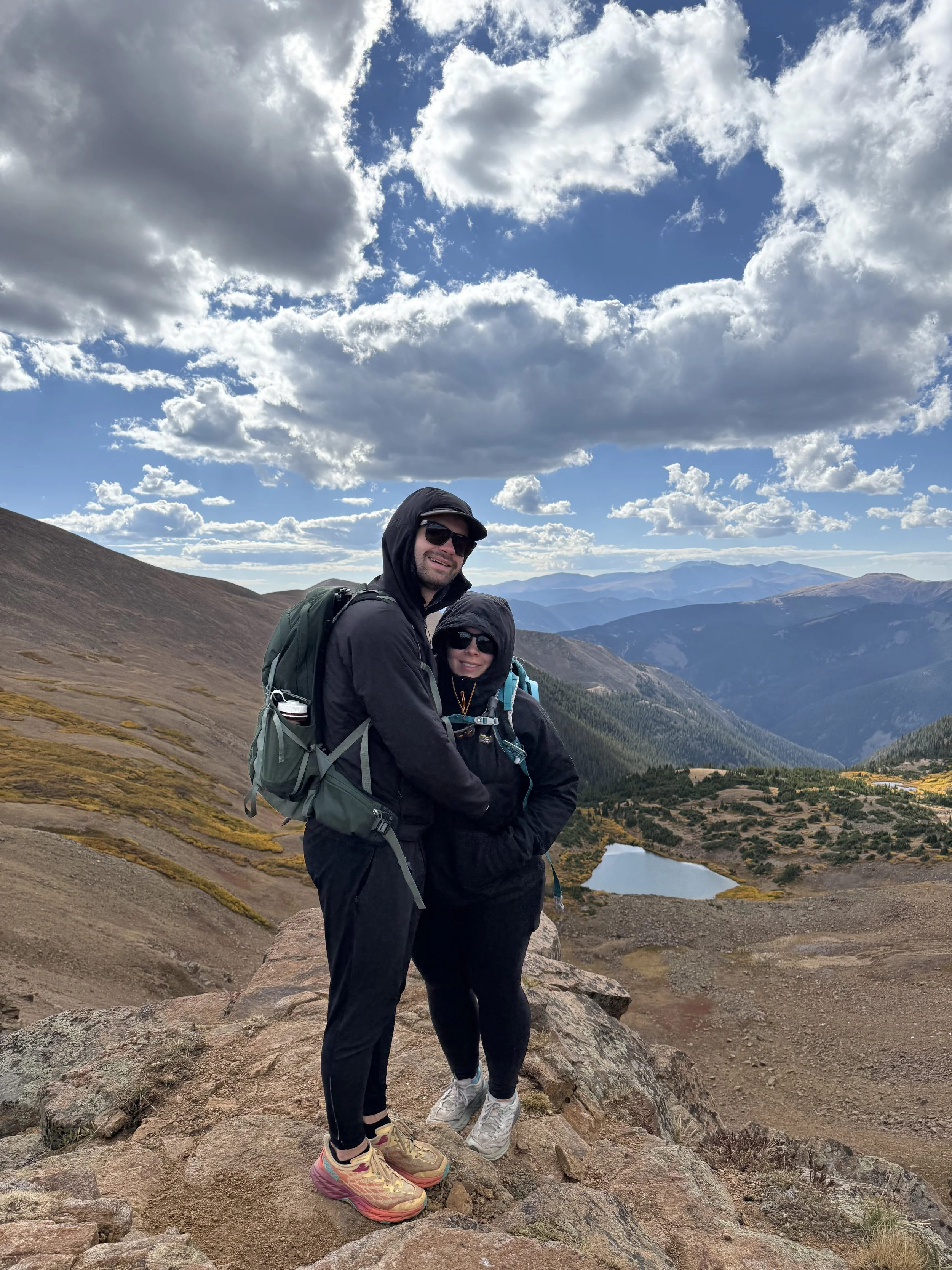 A man and woman in black hiking gear standing on rocky terrain in a mountainous landscape with a lake, trees, and distant mountains under a partly cloudy sky.