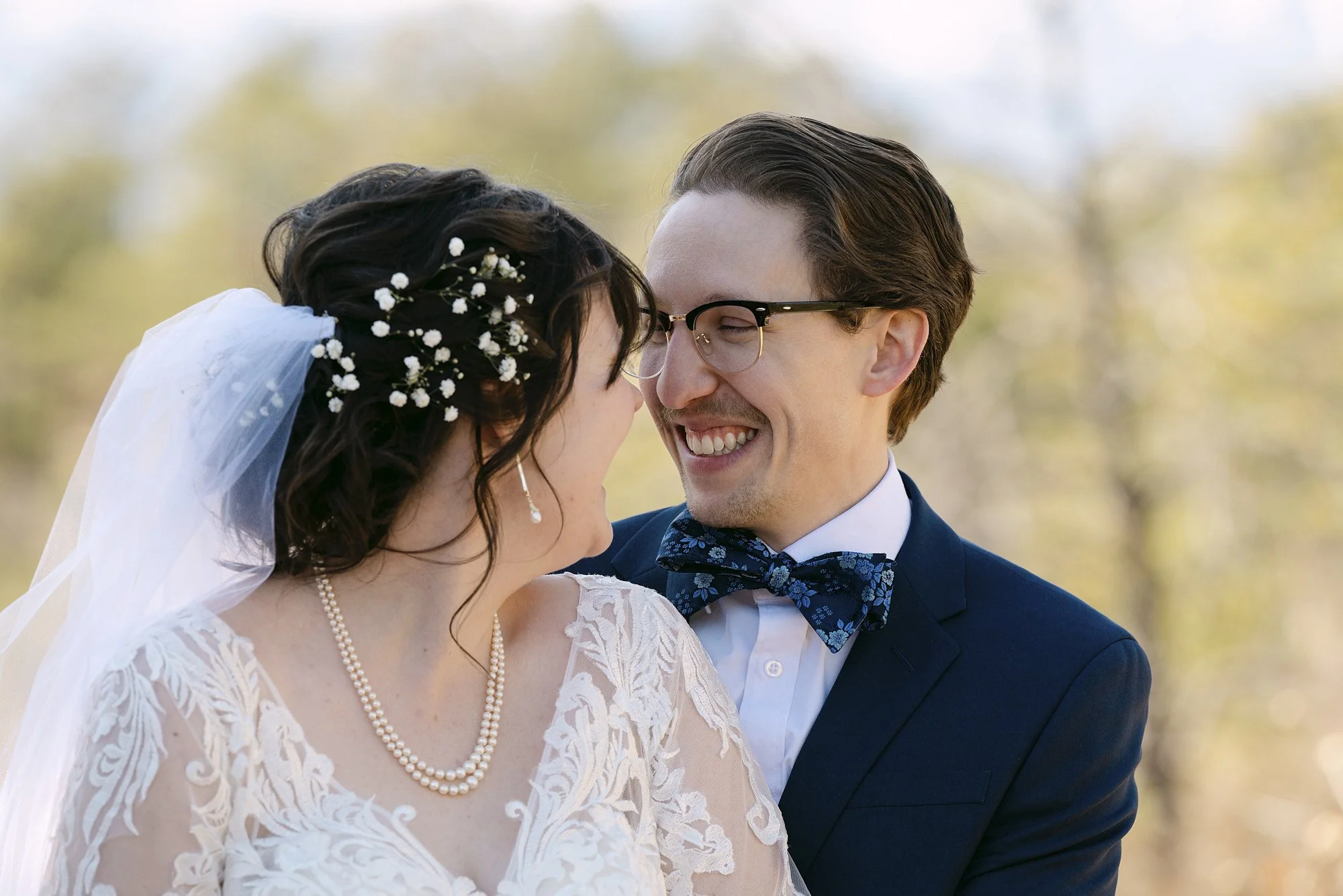 A smiling bride and groom close together outdoors, with a blurred background of trees, dressed in wedding attire.