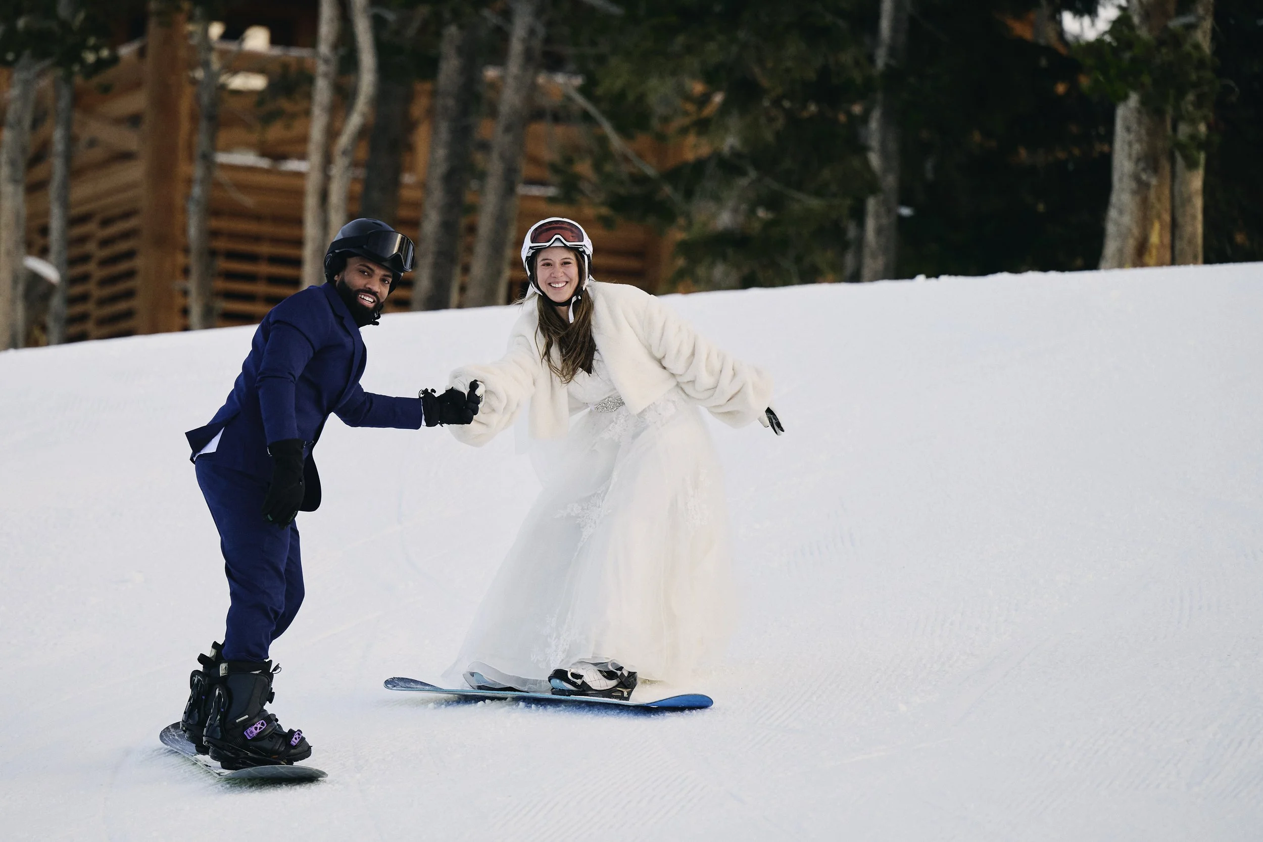 A man and woman in wedding attire snowboarding on a snowy slope, holding hands and smiling.