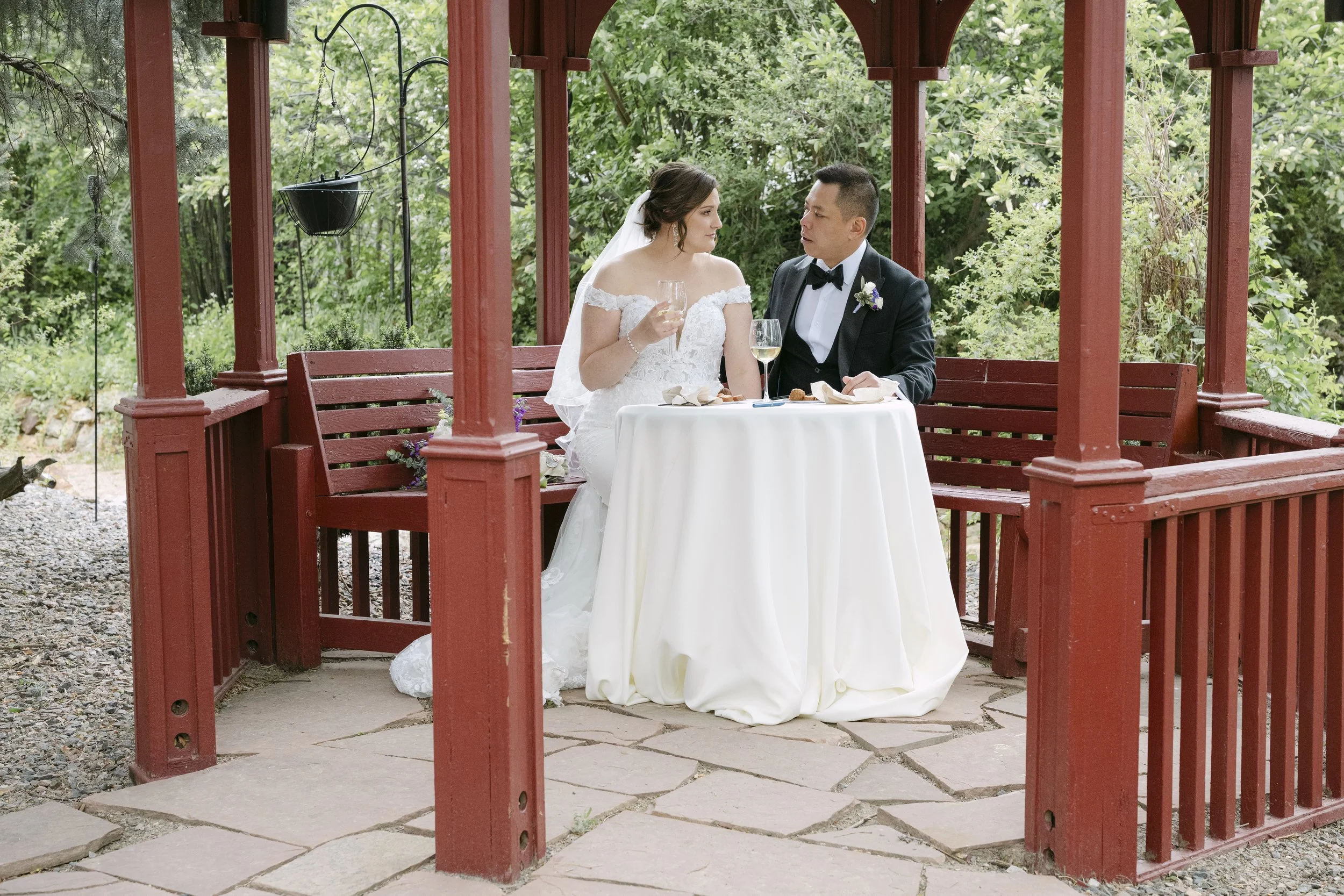Bride and groom sitting together on a red wooden gazebo, surrounded by green trees, with the bride holding a glass of white wine and the groom looking at her, during a wedding celebration.