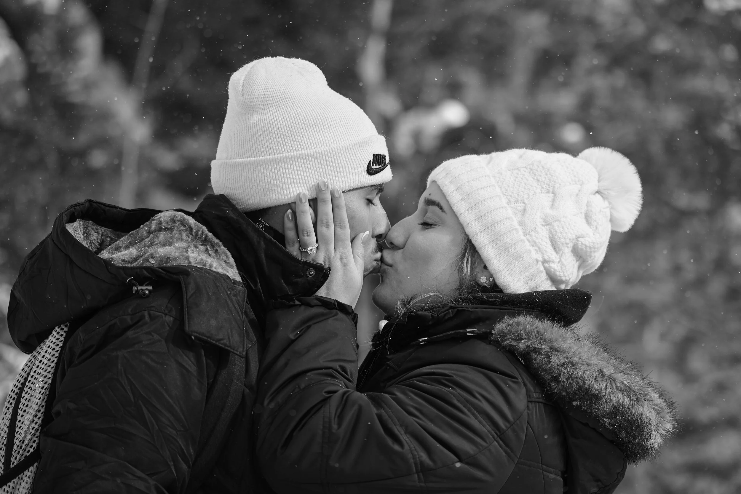 A couple in winter clothes sharing a kiss outdoors, with snow falling around them.