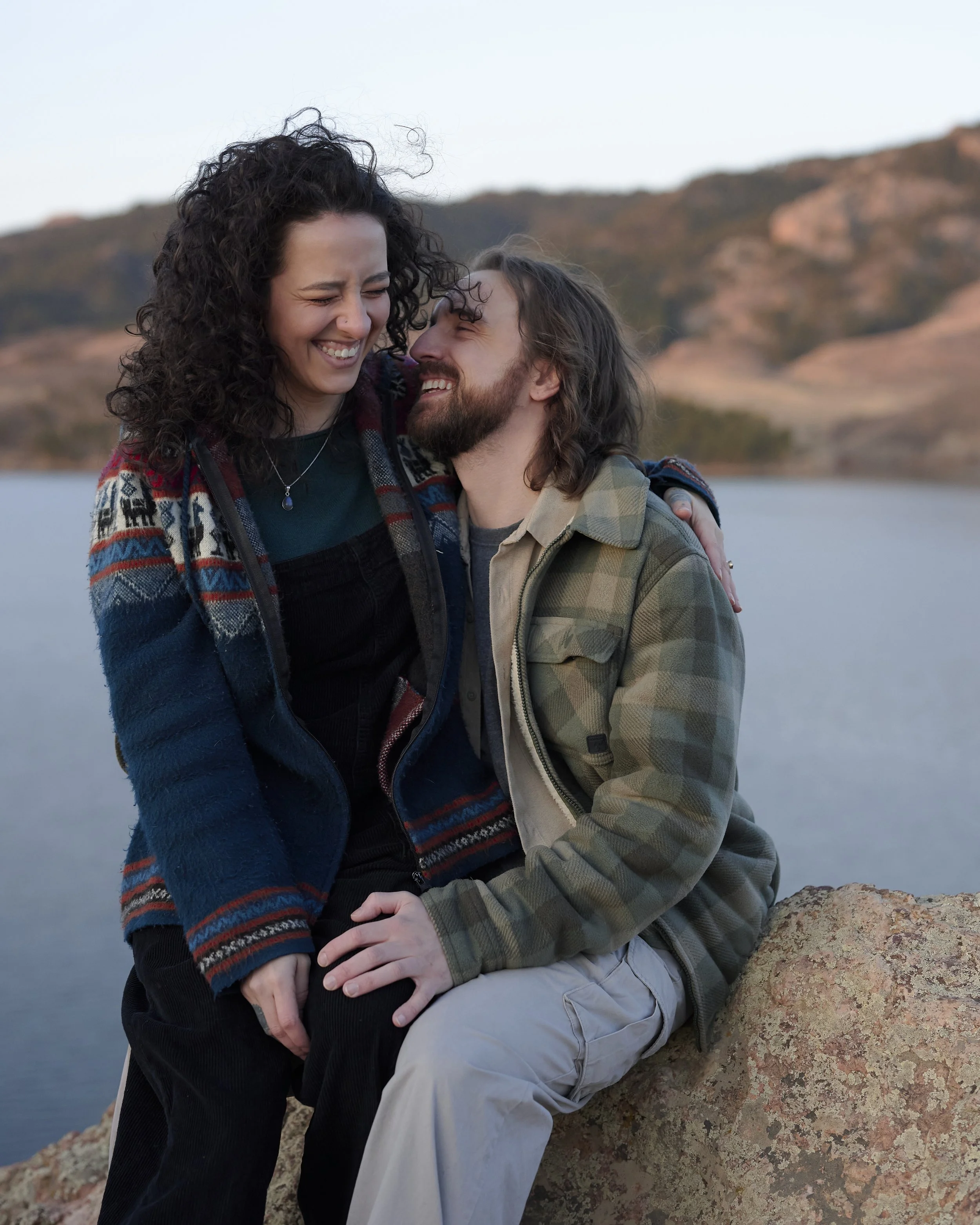 A couple sitting on a rock near a body of water, smiling and embracing each other, with mountains in the background.