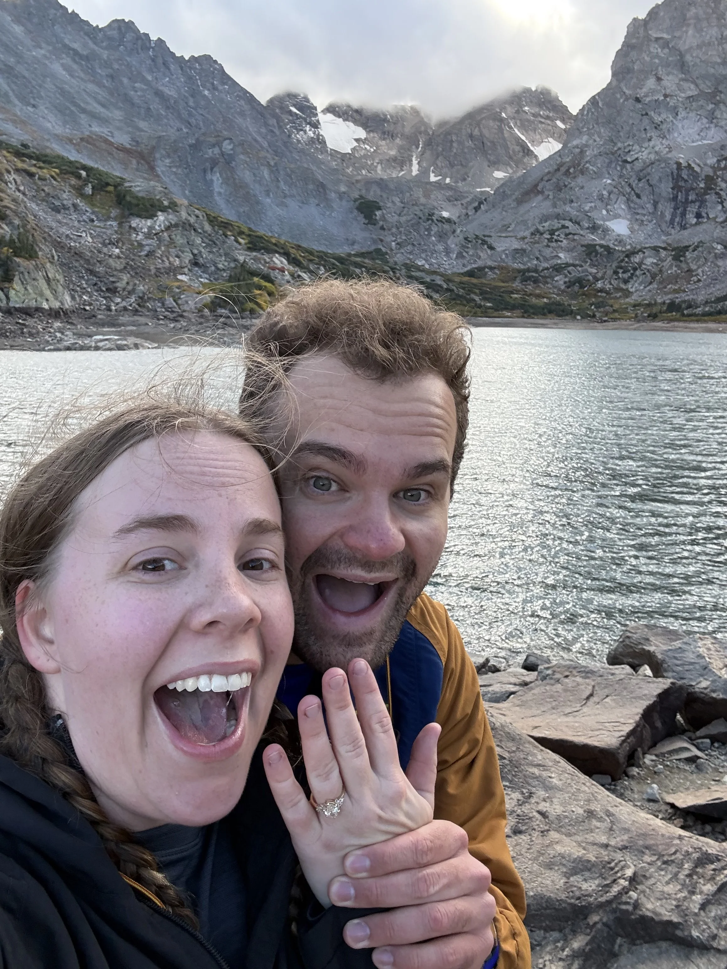 Two people, a woman and a man, are taking a selfie near a mountain lake. The woman shows her engagement ring, and they are both smiling excitedly. Behind them are rocky shores, water, and towering mountains with some snow patches and clouds in the sky.