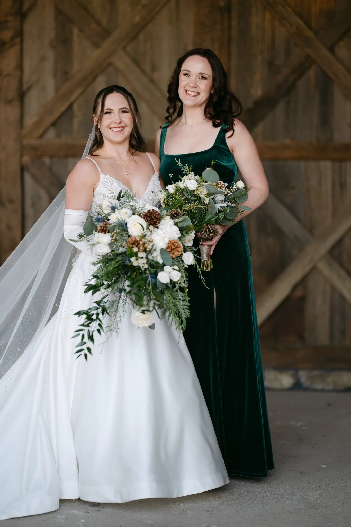 Two women smiling and holding bouquets, one in a wedding dress and the other in a dark green dress, standing in front of a rustic wooden background.