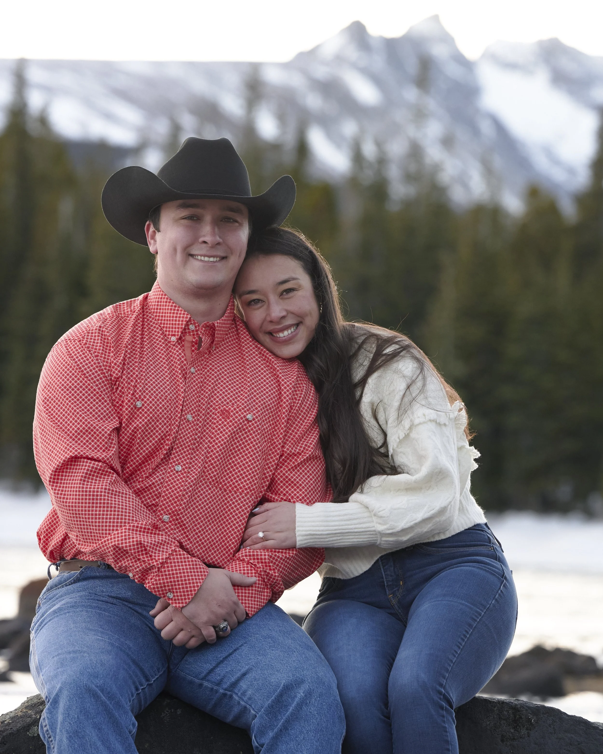 A smiling couple sitting outdoors in front of a mountain landscape, with the man wearing a cowboy hat and the woman hugging him.