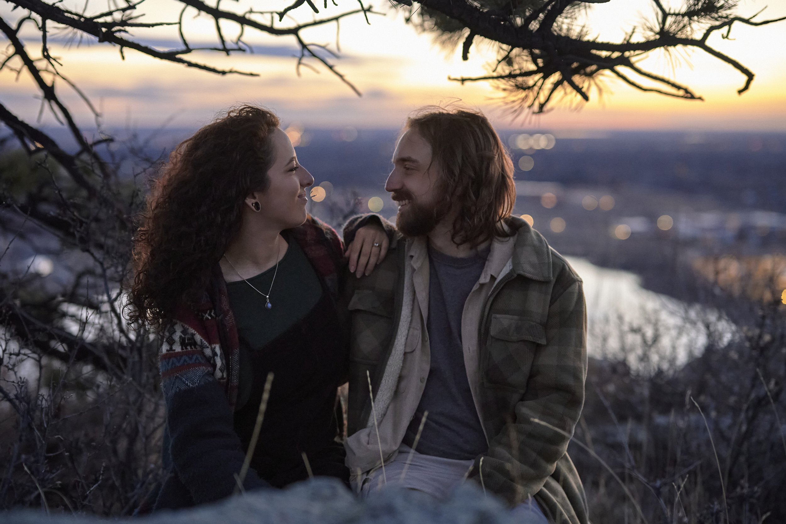 A couple sitting outdoors at sunset, smiling and looking at each other, with a scenic river and city lights in the background.