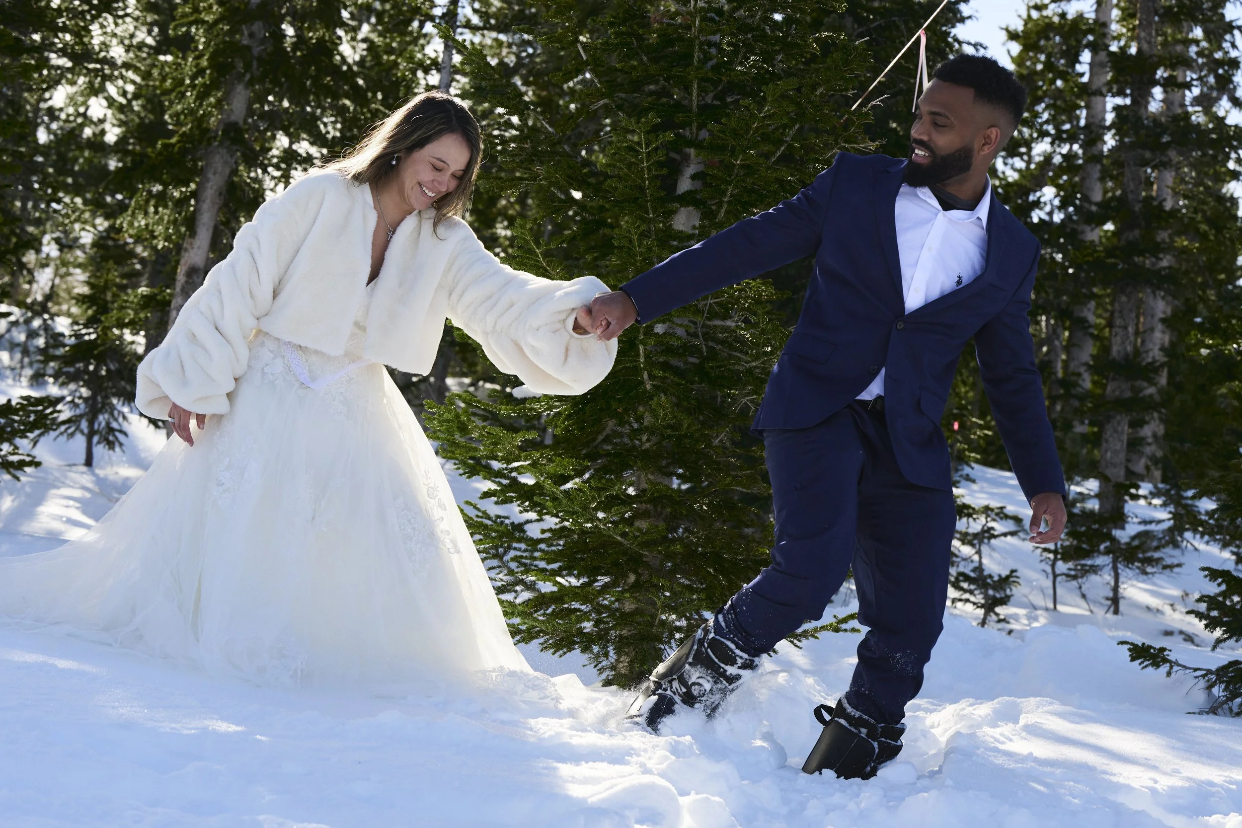 A couple dressed in wedding attire having fun together in the snow in a forest, with trees and snow-covered ground visible.