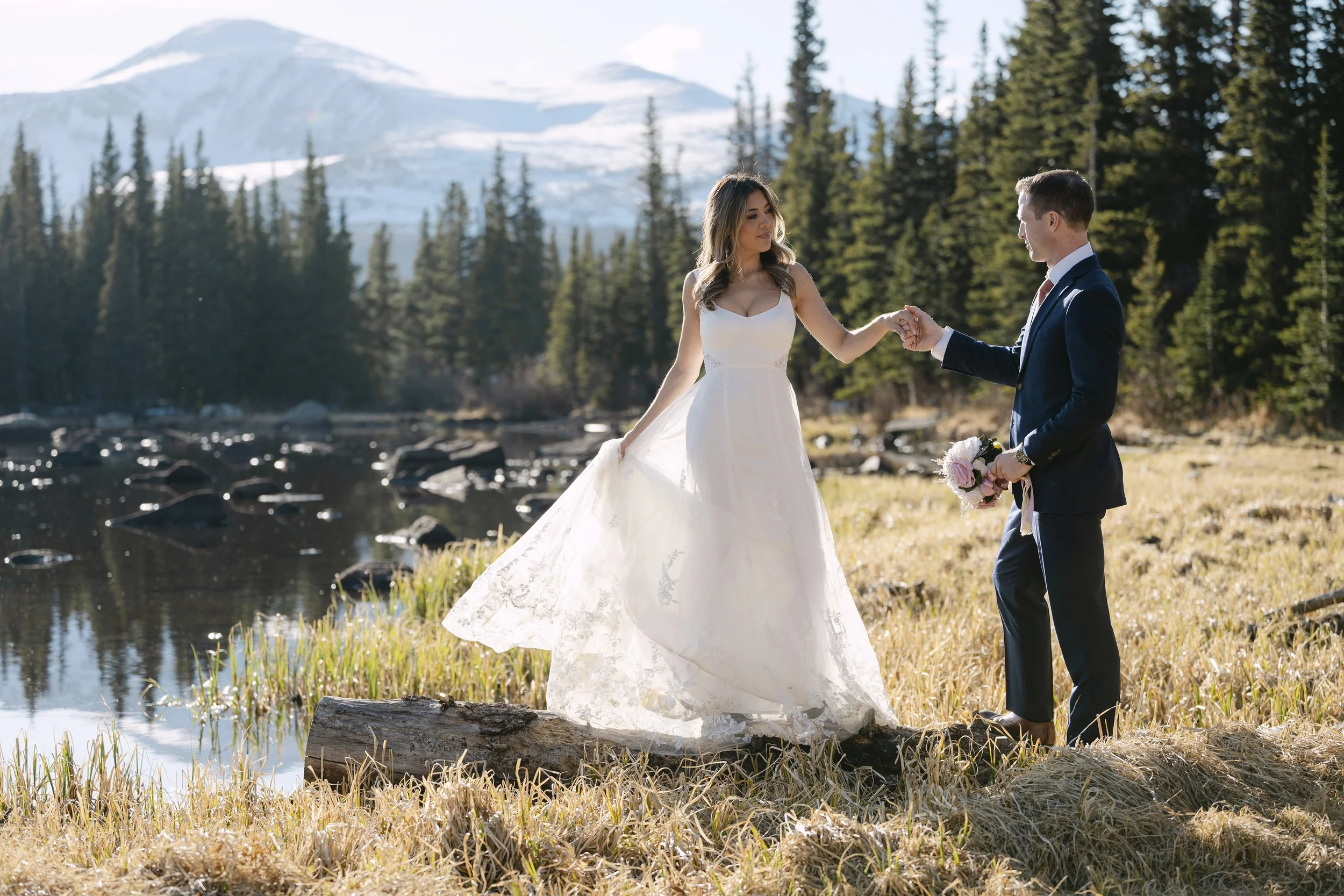 A bride rests her hand in  her groom's hand at the edge of Red Rock Lake with the snow-covered Indian Peaks glowing in golden hour light in the background