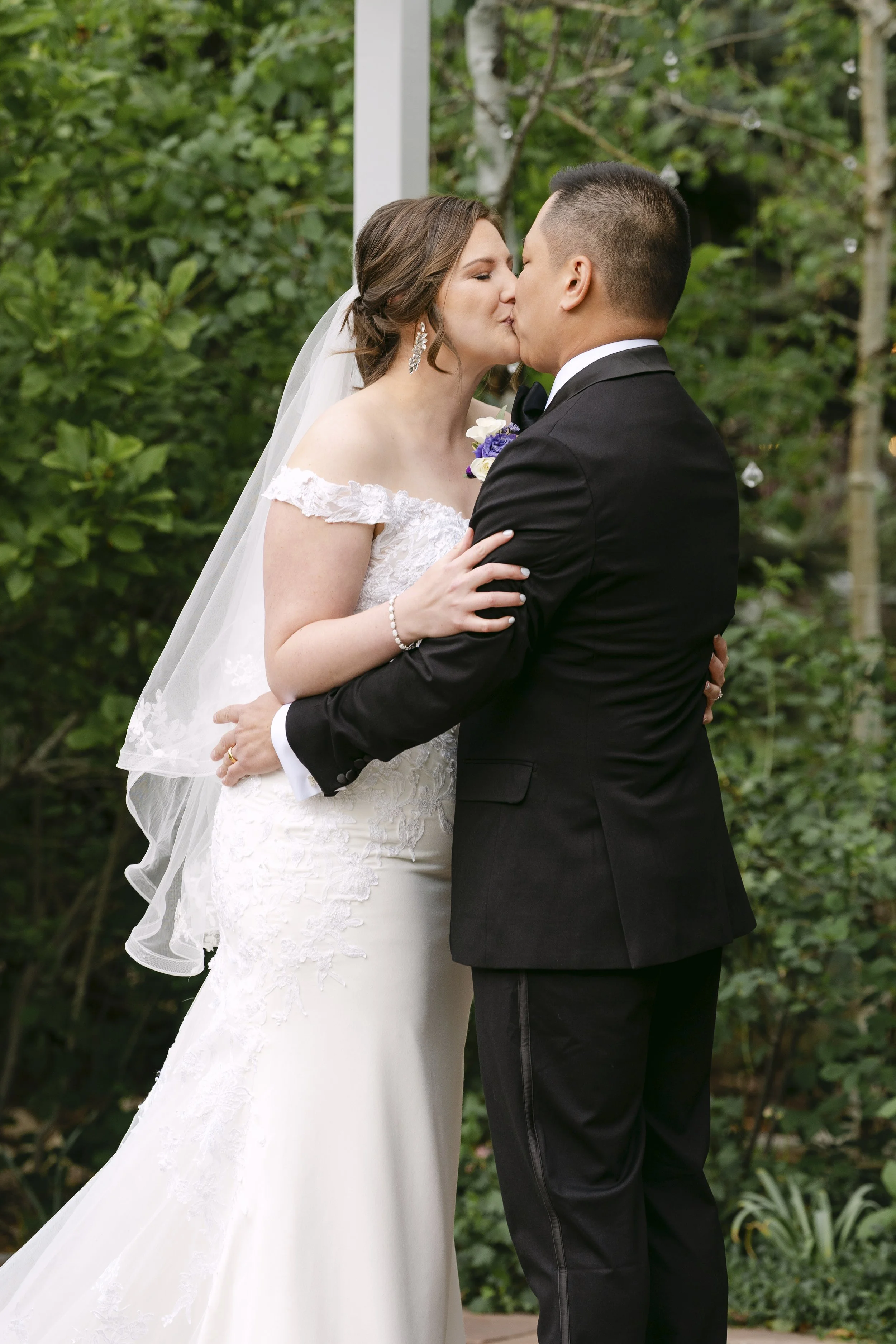 Bride and groom sharing a kiss outdoors, surrounded by greenery, during their wedding ceremony.