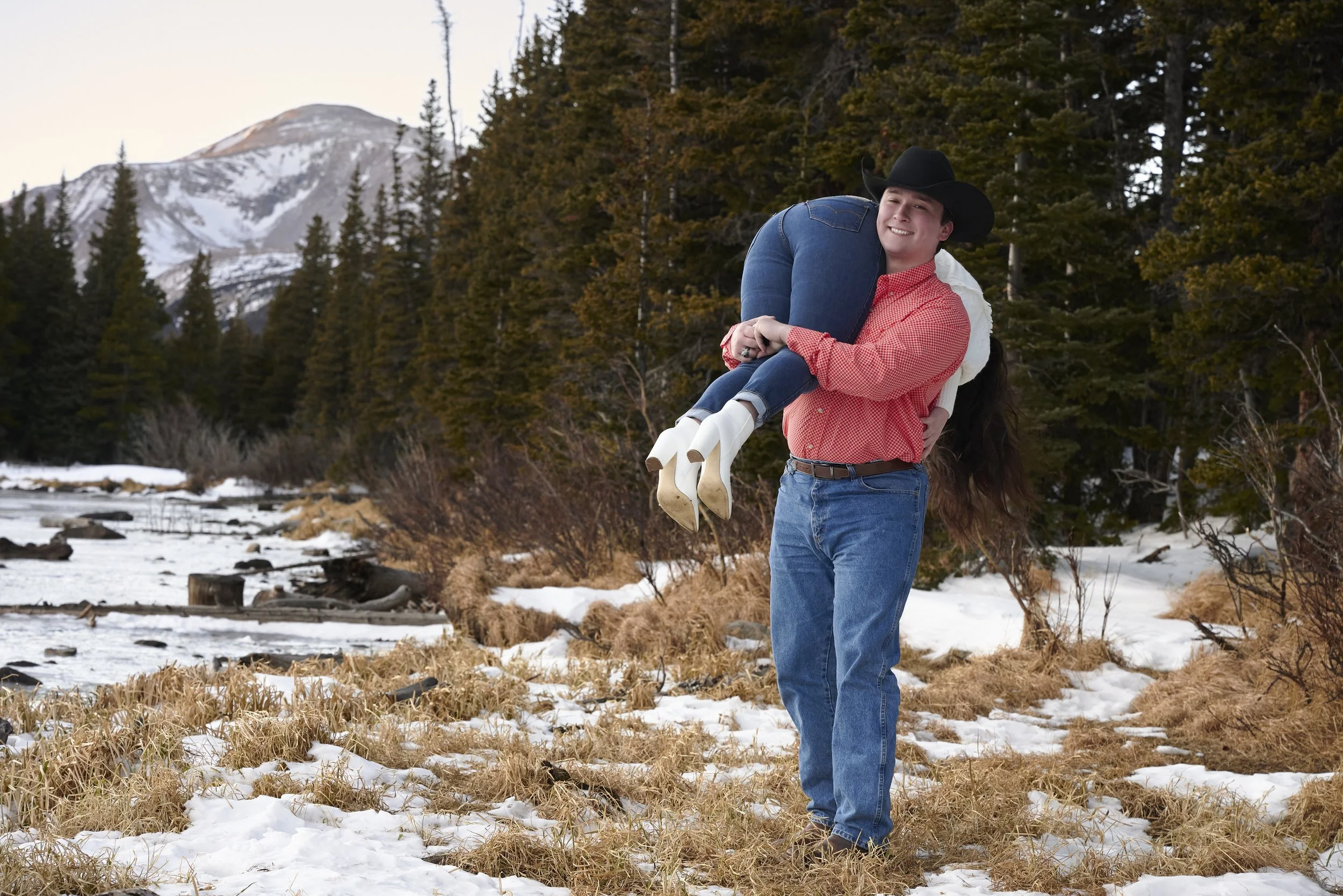 A man wearing a cowboy hat, red checkered shirt, and jeans holding a woman in a white jacket and blue jeans, outdoors in a snowy, wooded landscape with mountains in the background.