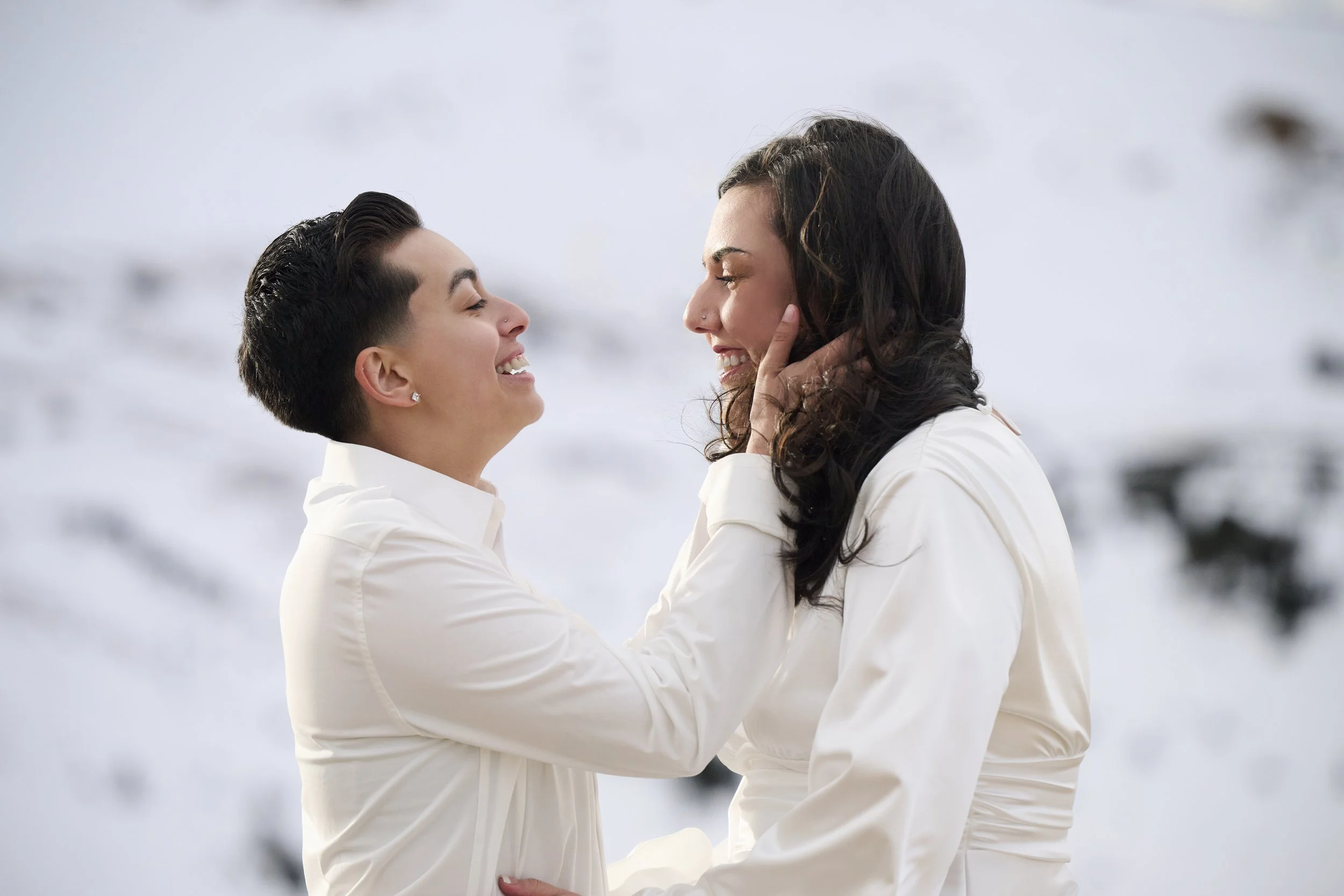 Two women in white outfits smiling and embracing each other outdoors with a snow-covered landscape in the background.