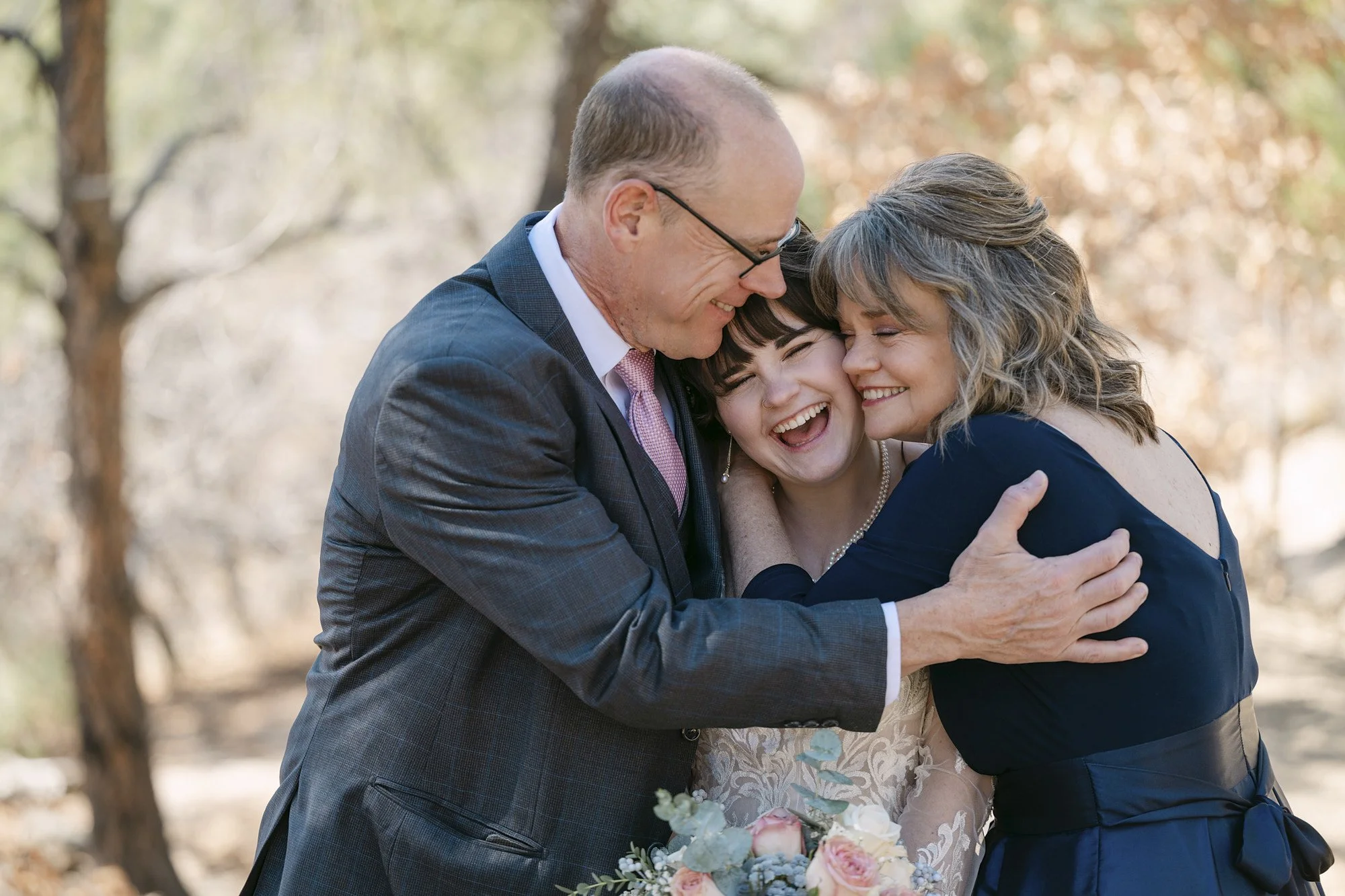 Three people, a man, a woman, and a young woman, are happily hugging outdoors. The man and woman are older and the young woman has dark hair. They are smiling and appear joyful. The woman holds a bouquet of flowers.