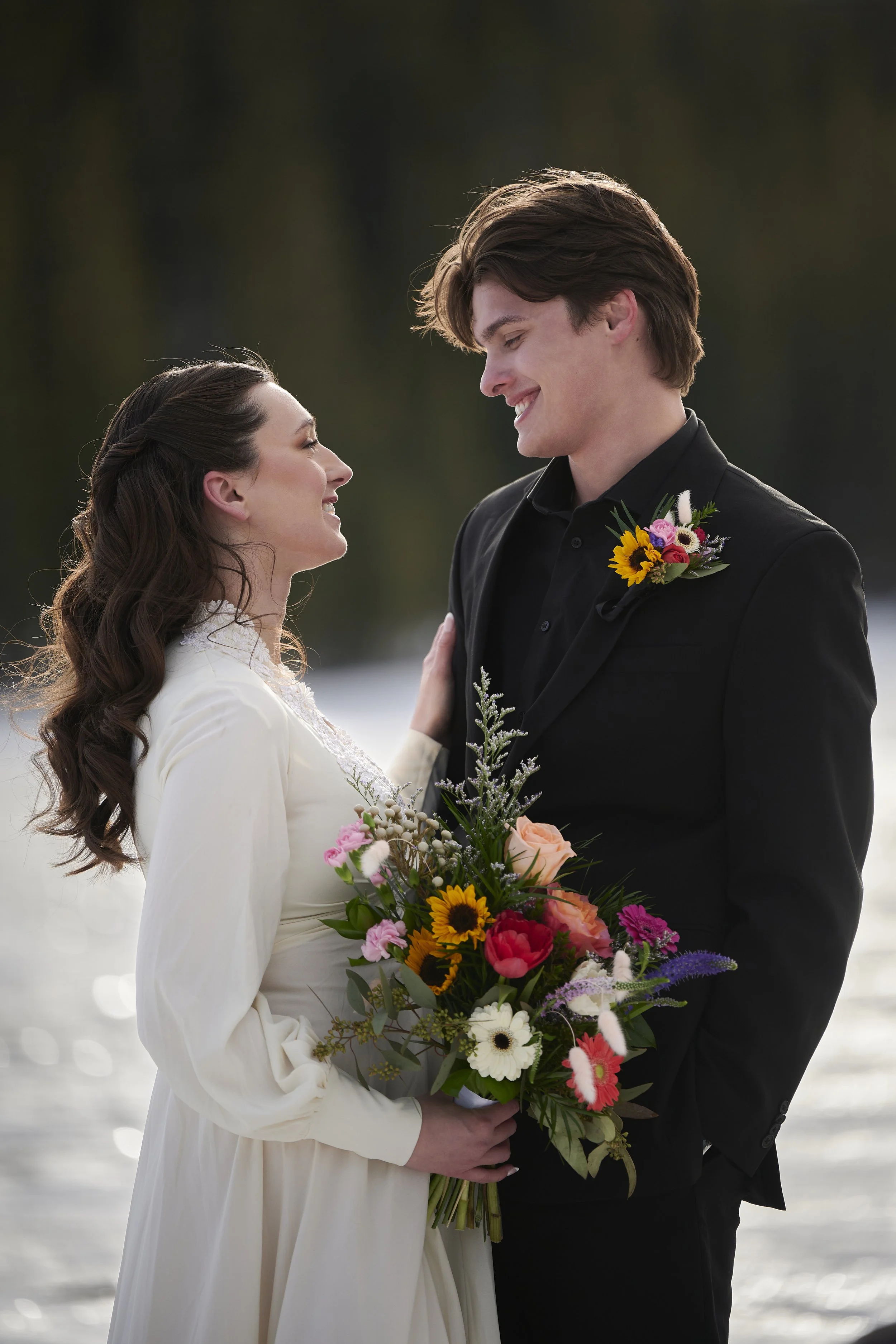 A bride and groom on their wedding day, smiling at each other outdoors near a body of water, with the bride holding a colorful bouquet and the groom wearing a boutonniere.