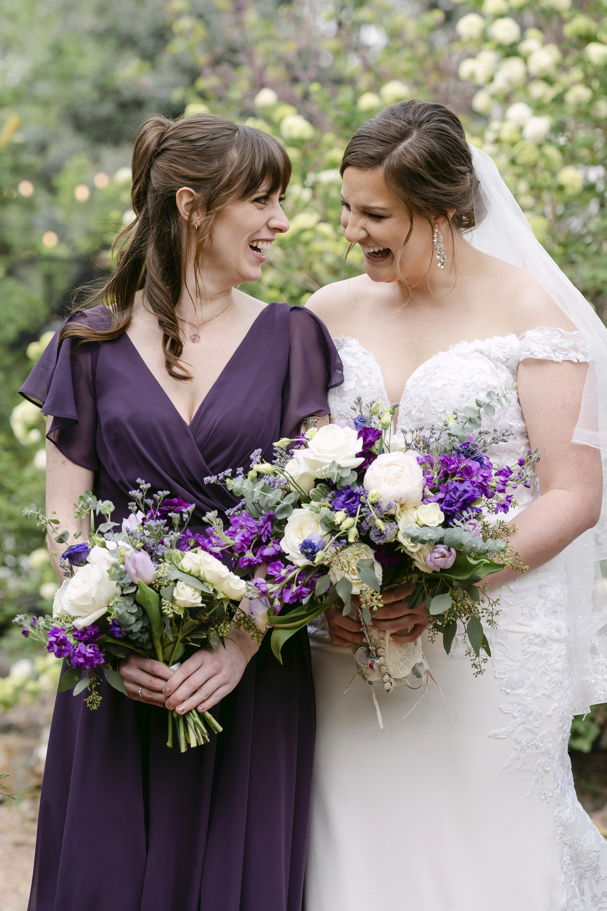 A bride in a white lace wedding dress holding a bouquet of purple, white, and green flowers, standing next to a woman in a purple dress, both smiling and looking at each other outdoors with greenery and blooming roses in the background.
