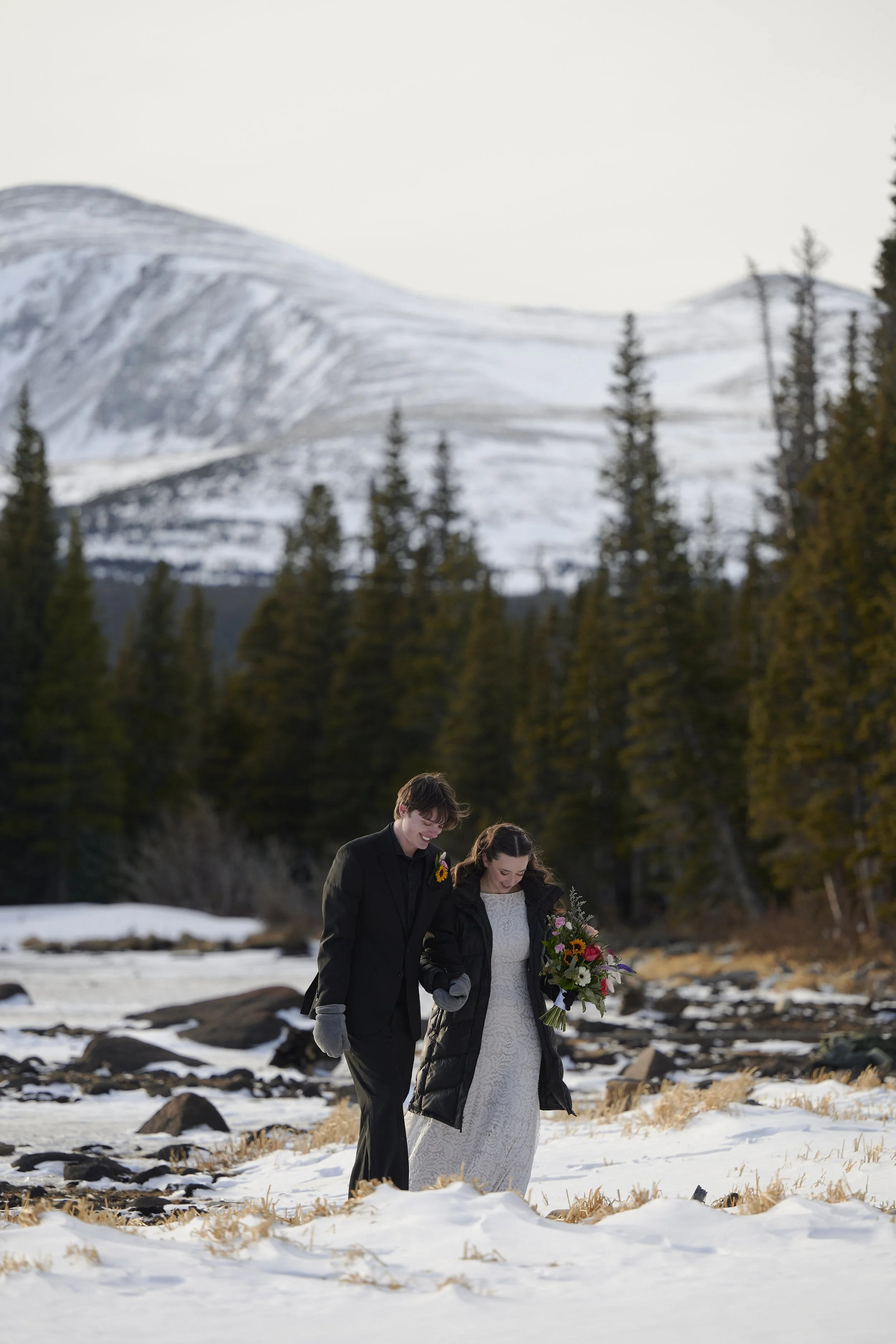 A bride and groom walking hand in hand outdoors on snow-covered ground, with a mountain and pine trees in the background. The bride holds a bouquet of colorful flowers, and they are dressed warmly.