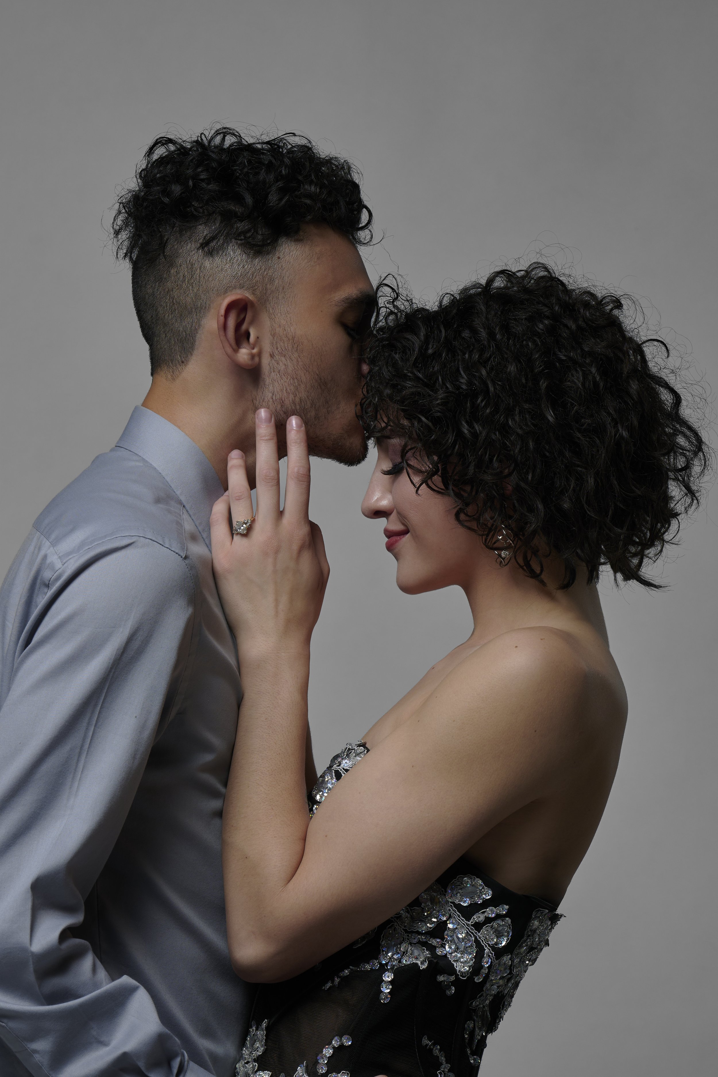 A man and woman with curly hair sharing a kiss, with the man's hand gently touching the woman's face, against a neutral gray background.