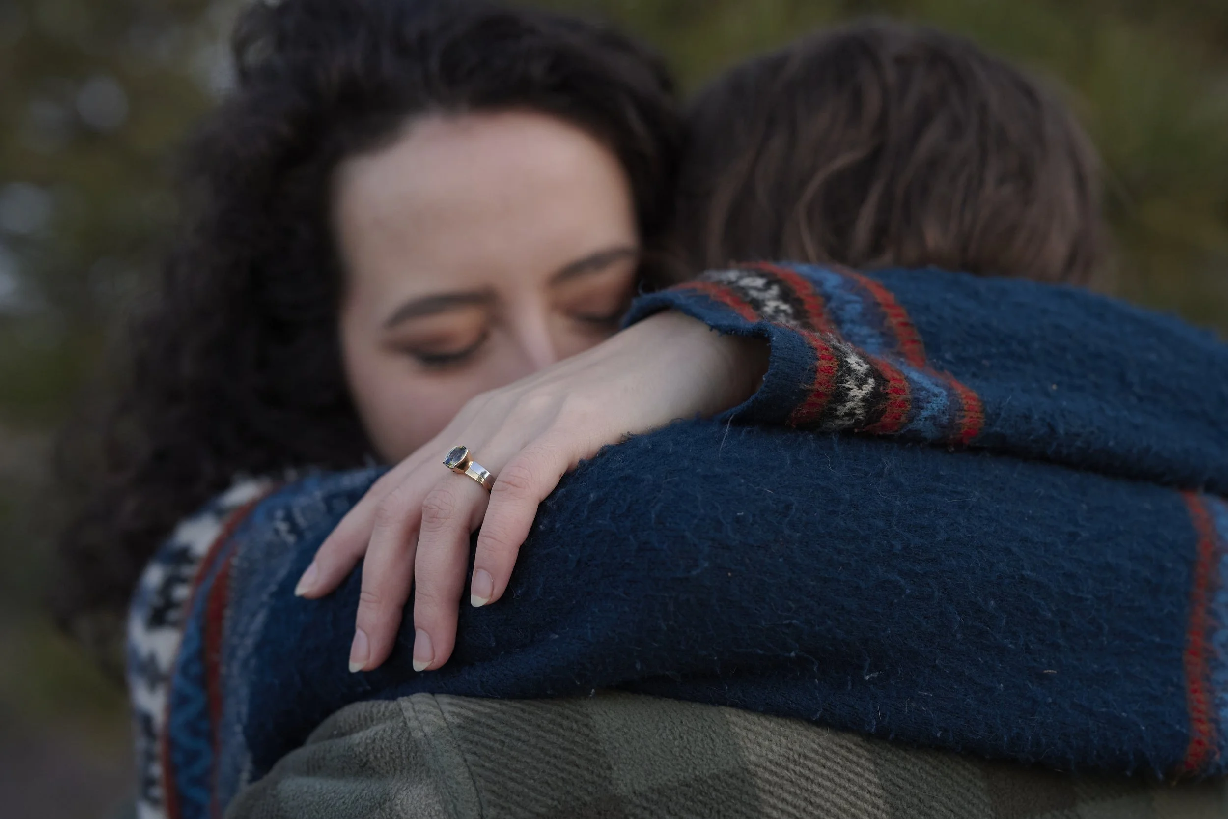 A woman with dark, curly hair and makeup is hugging a person, with her eyes closed and an emotional expression. She is wearing a gold ring with a dark stone on her finger and a blue, red, and black patterned wool sweater.