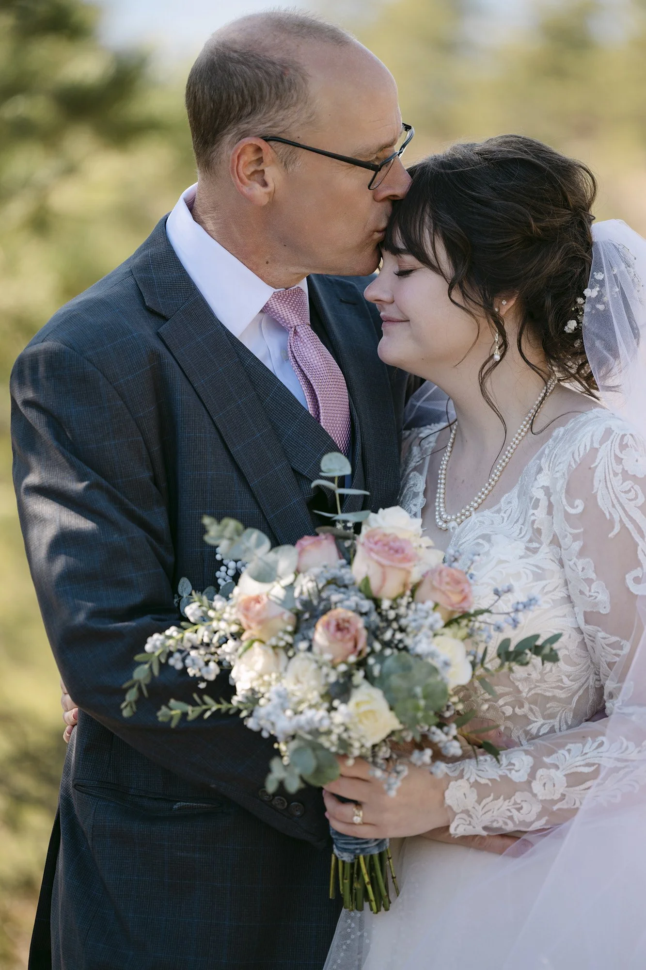 A father kissing his daughter on the forehead during a wedding, holding a bouquet of pink and white flowers with greenery.