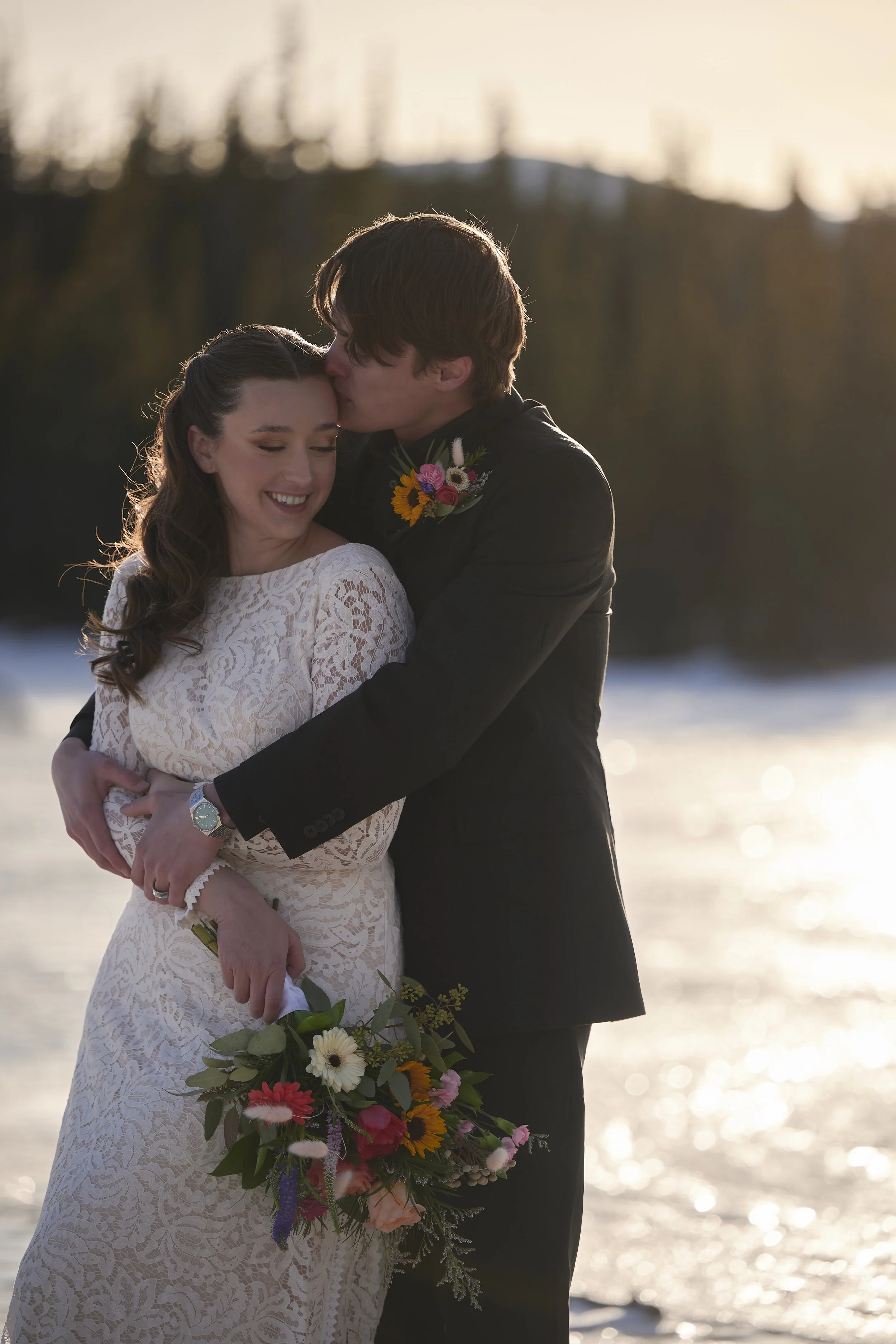 A bride and groom in an embrace on a snowy landscape at sunset, with the groom kissing the bride's forehead. The bride is holding a colorful bouquet, and they are both smiling.