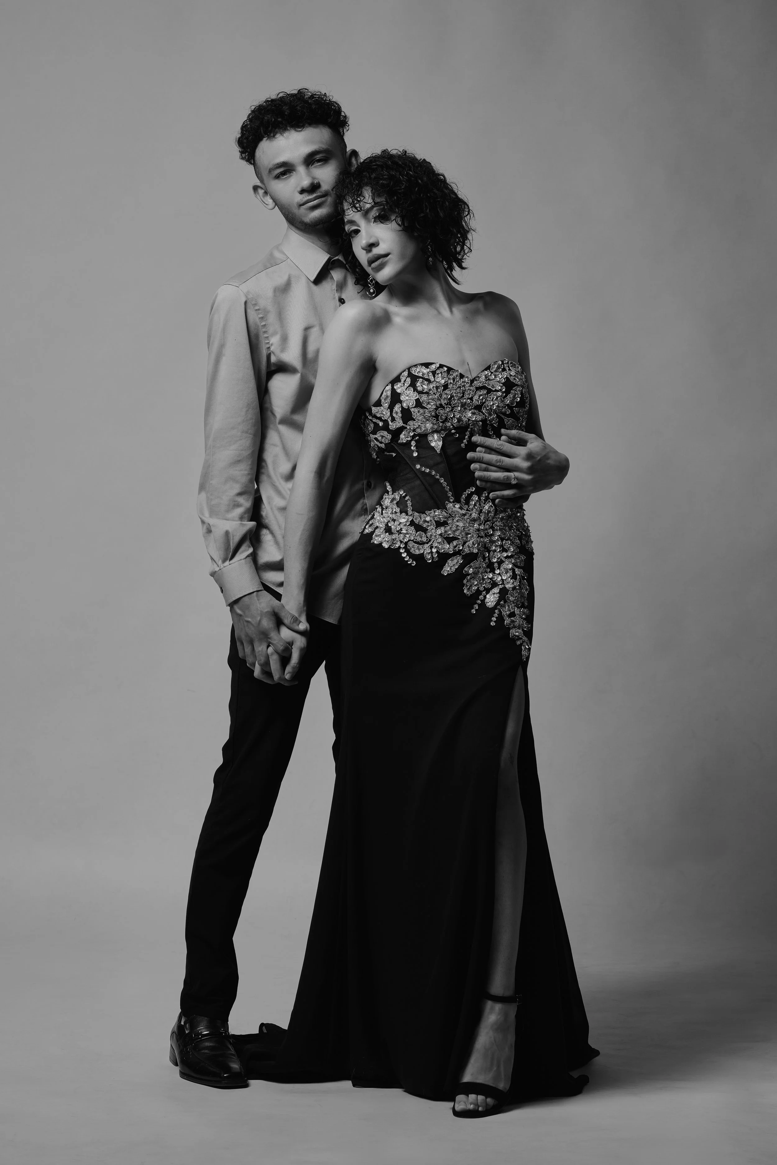A black and white photograph of a man and woman standing close, holding hands. The woman wears a strapless, embellished gown with a slit, and the man wears a button-up shirt and dark trousers. They have serious expressions and are posed against a pla