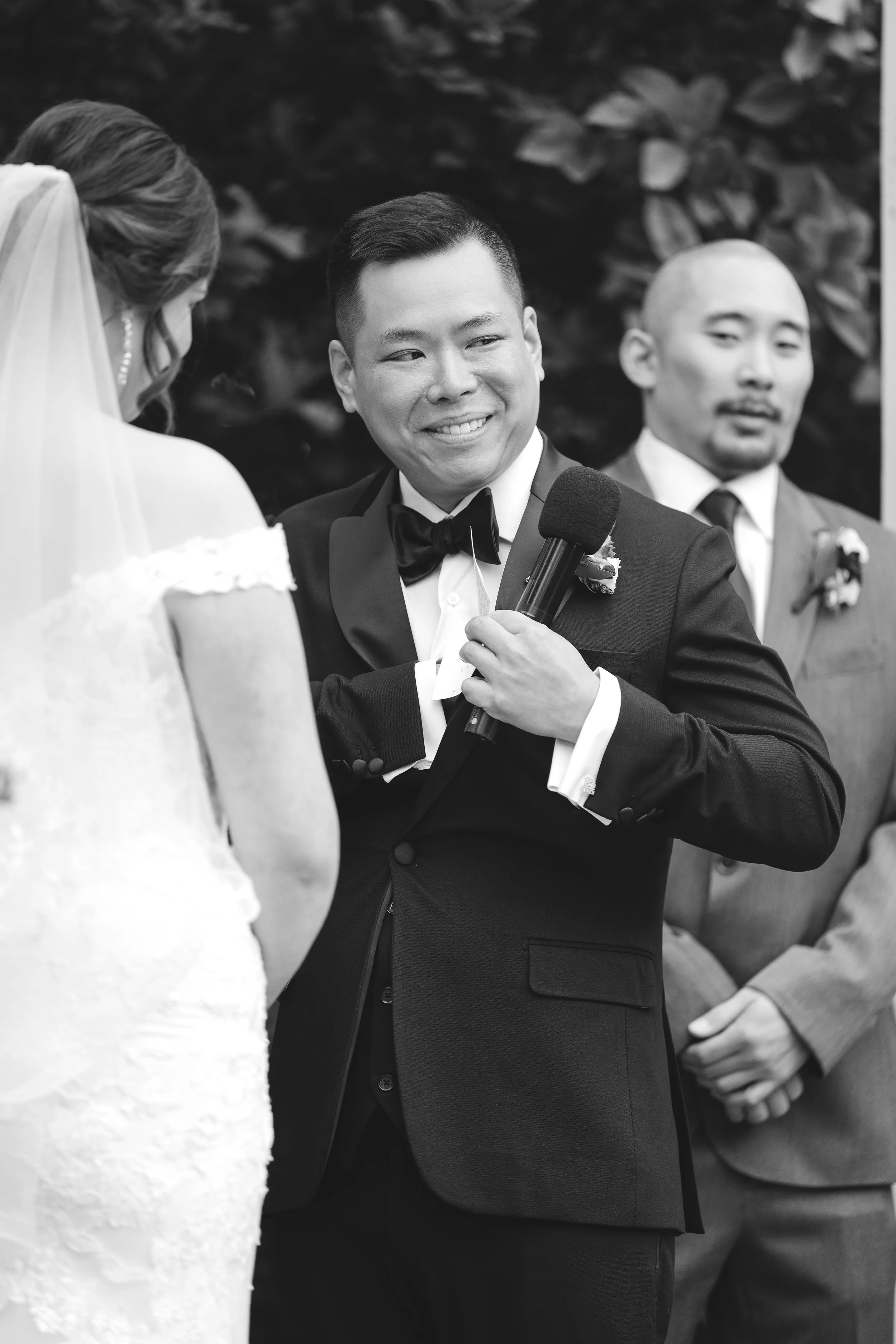 A black and white photo of a groom smiling and holding a microphone during a wedding ceremony, with a bride in a white dress and veil facing him, and another man in a suit in the background.
