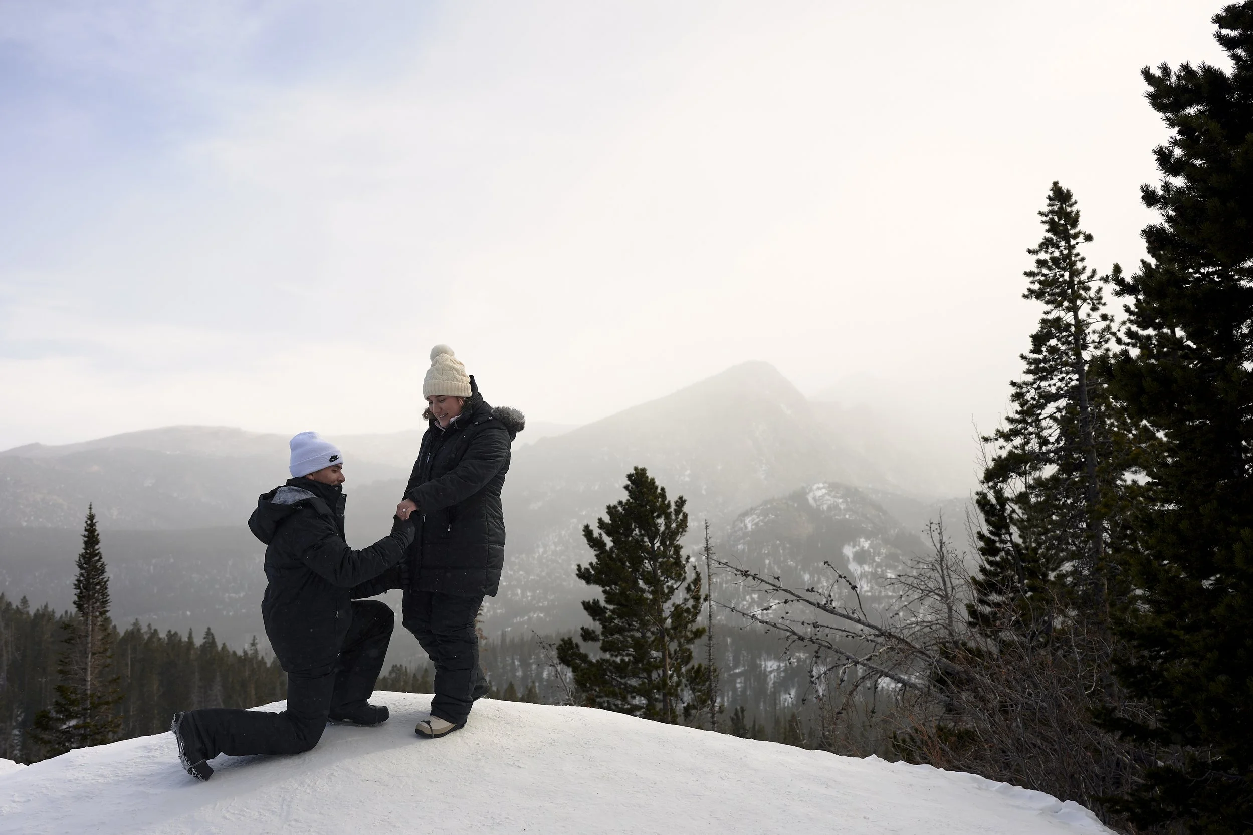 A man proposing marriage to a woman on snow-covered ground in a mountain landscape with pine trees and misty mountains in the background.
