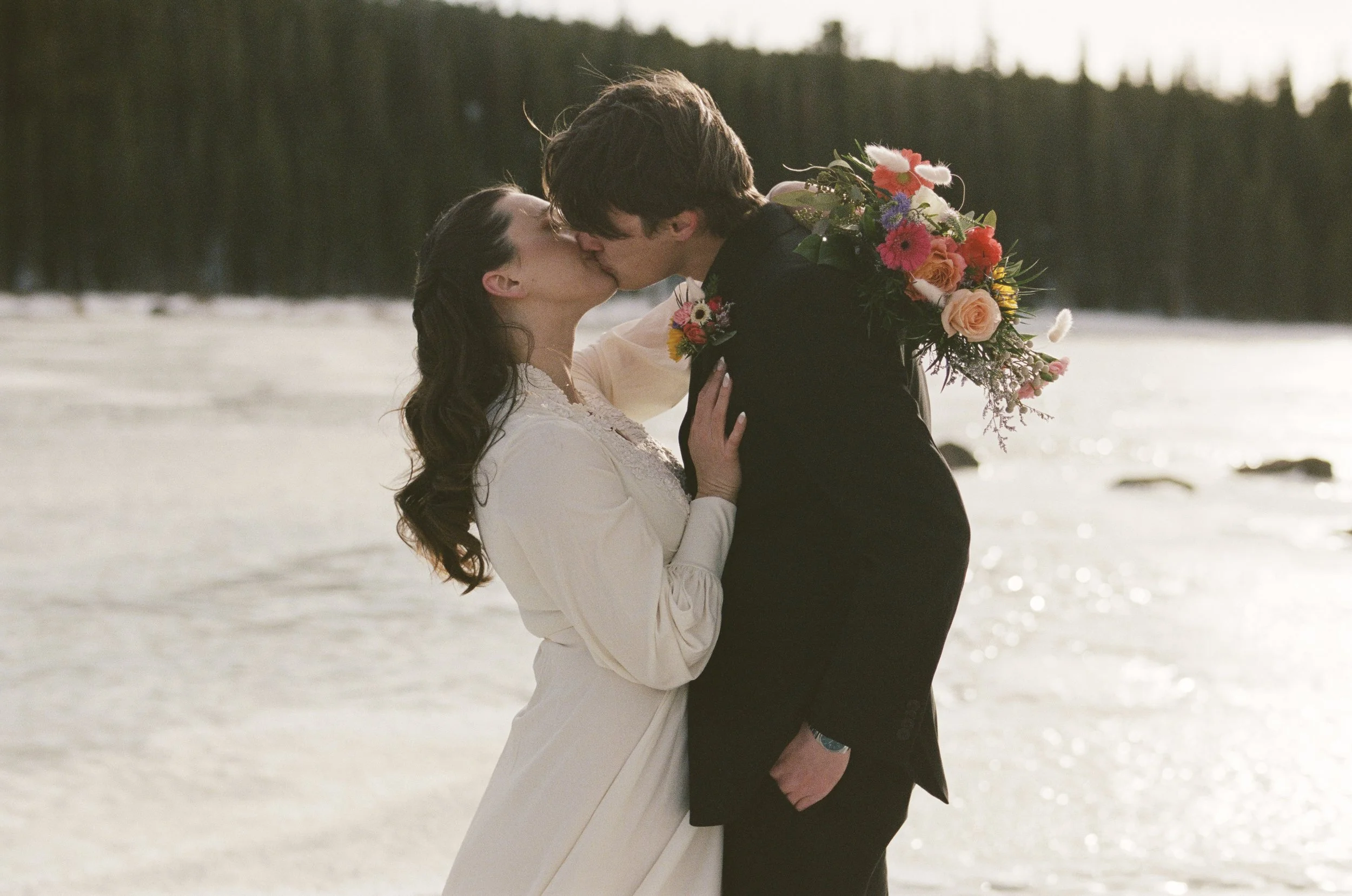 A couple kissing at the beach during sunset, the woman is wearing a white dress and the man is in a black suit, holding a colorful bouquet of flowers.