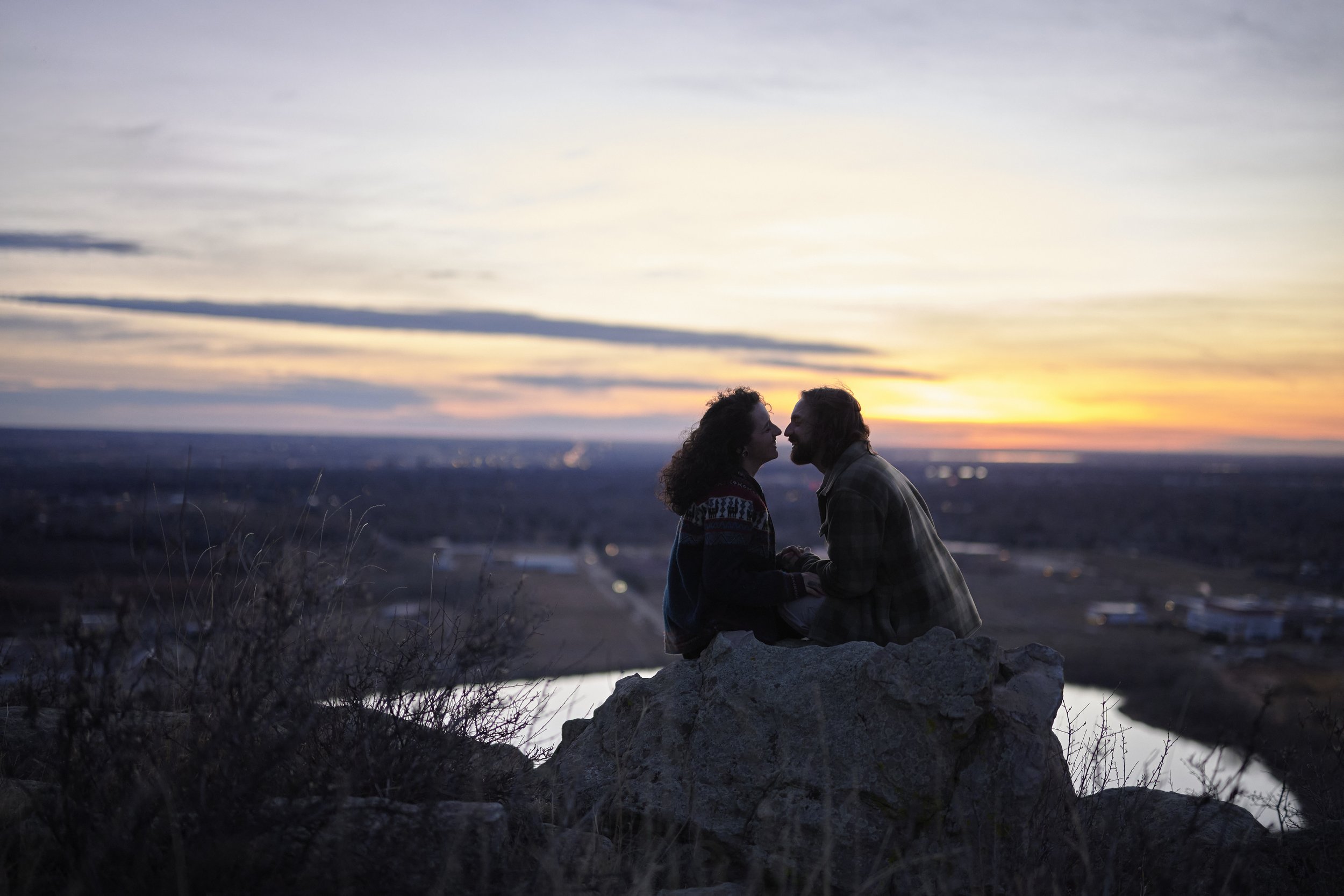 A couple sitting on a rock at sunset, facing each other and holding hands, with a scenic landscape and water body in the background.