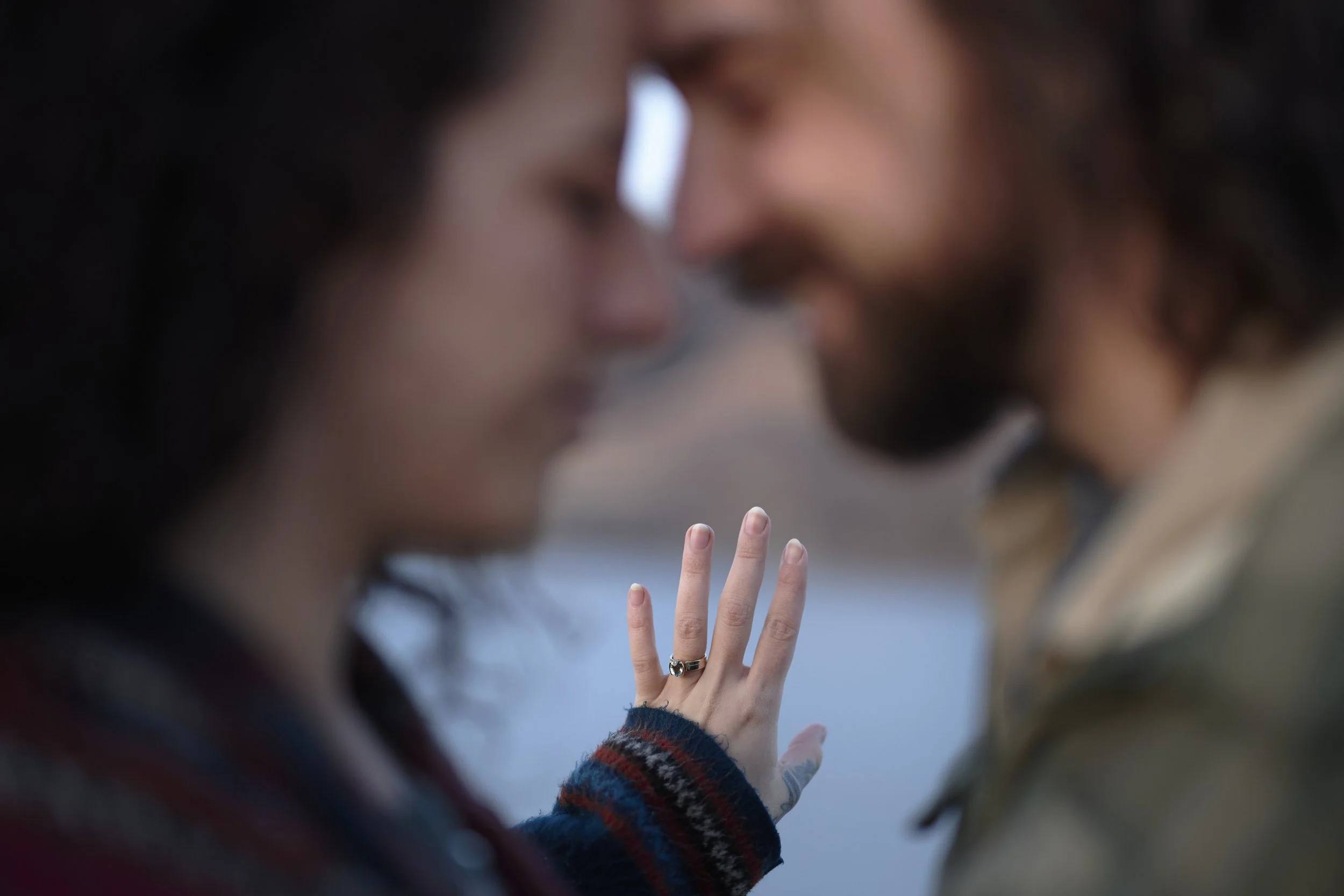 A couple with blurred faces in close proximity, with a woman's hand showing an engagement ring, near their foreheads.