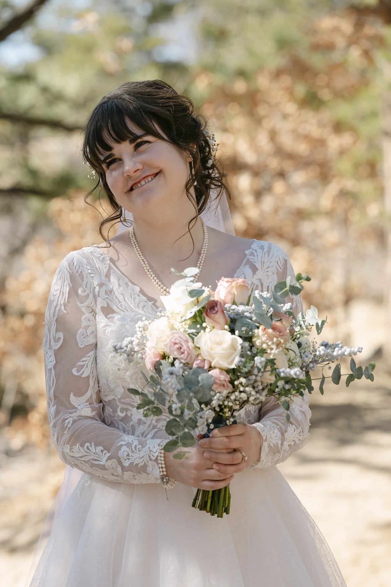 A smiling bride holding a bouquet of pink and white roses with greenery, standing outdoors with trees in the background.