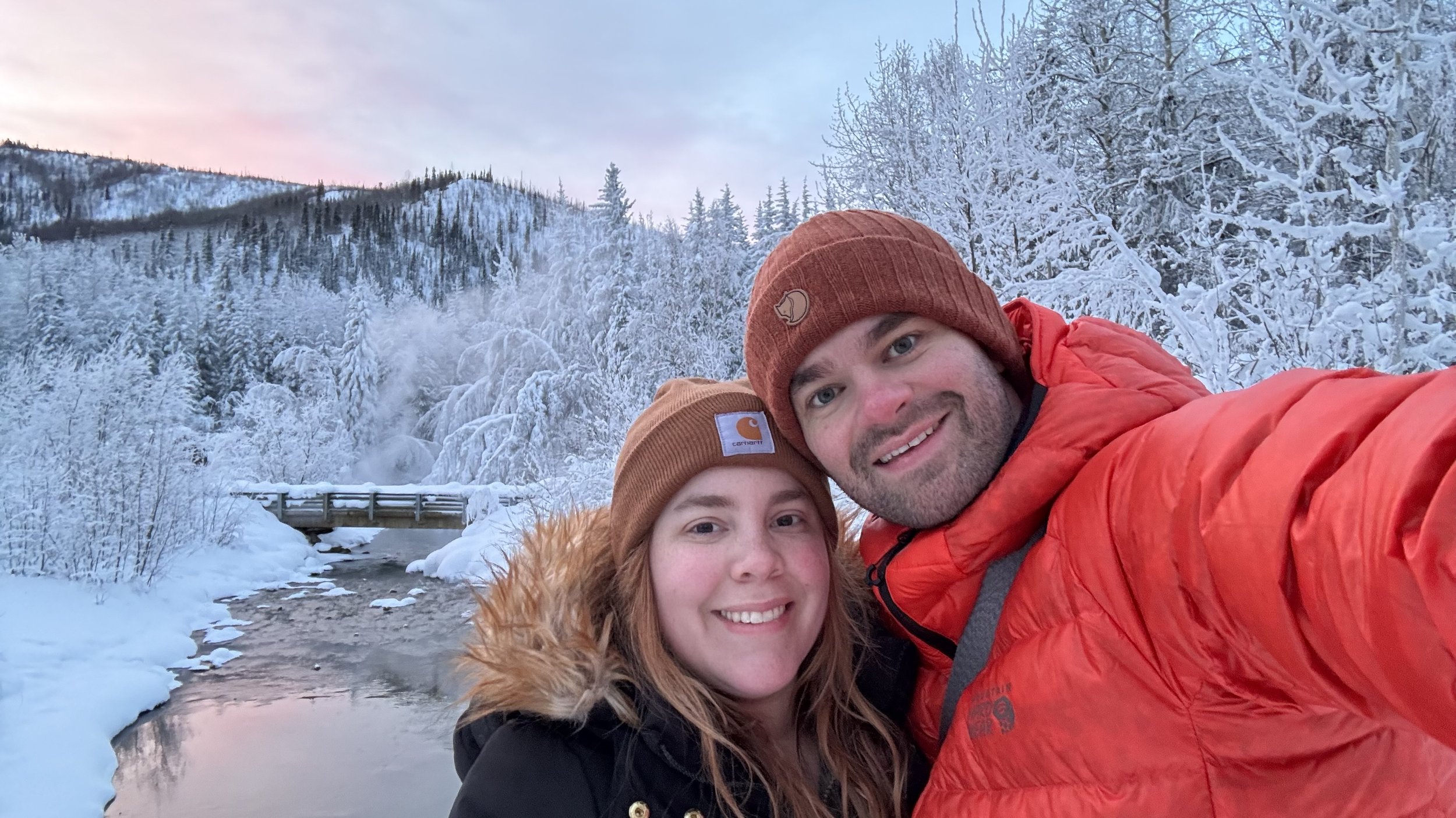 A smiling couple in winter clothing taking a selfie outdoors in a snowy, forested landscape with a small waterfall and snow-covered trees in the background.