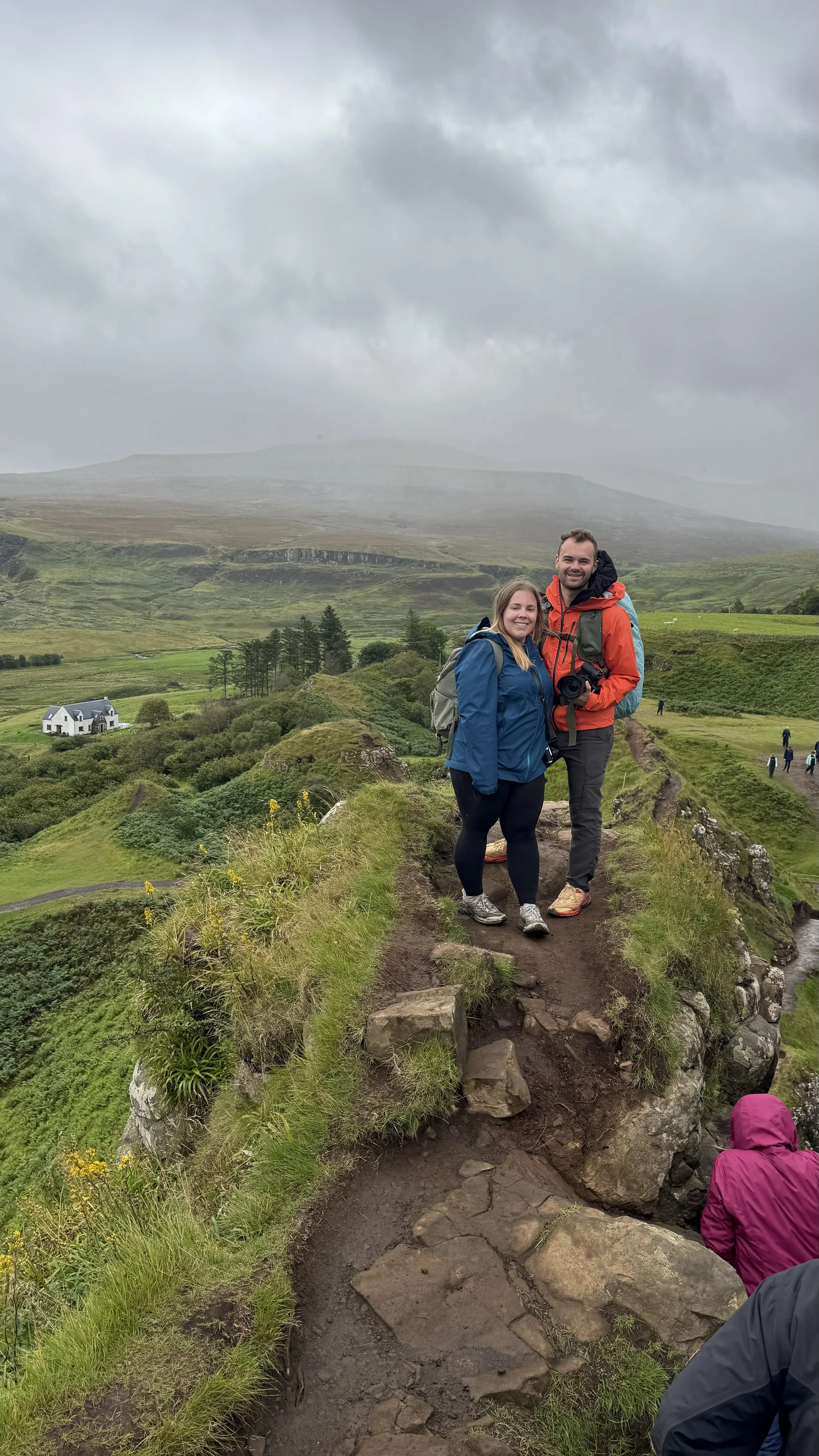 A man and woman in hiking gear standing on a trail atop a narrow ridge in a lush green landscape with a house and trees in the background, under cloudy skies.