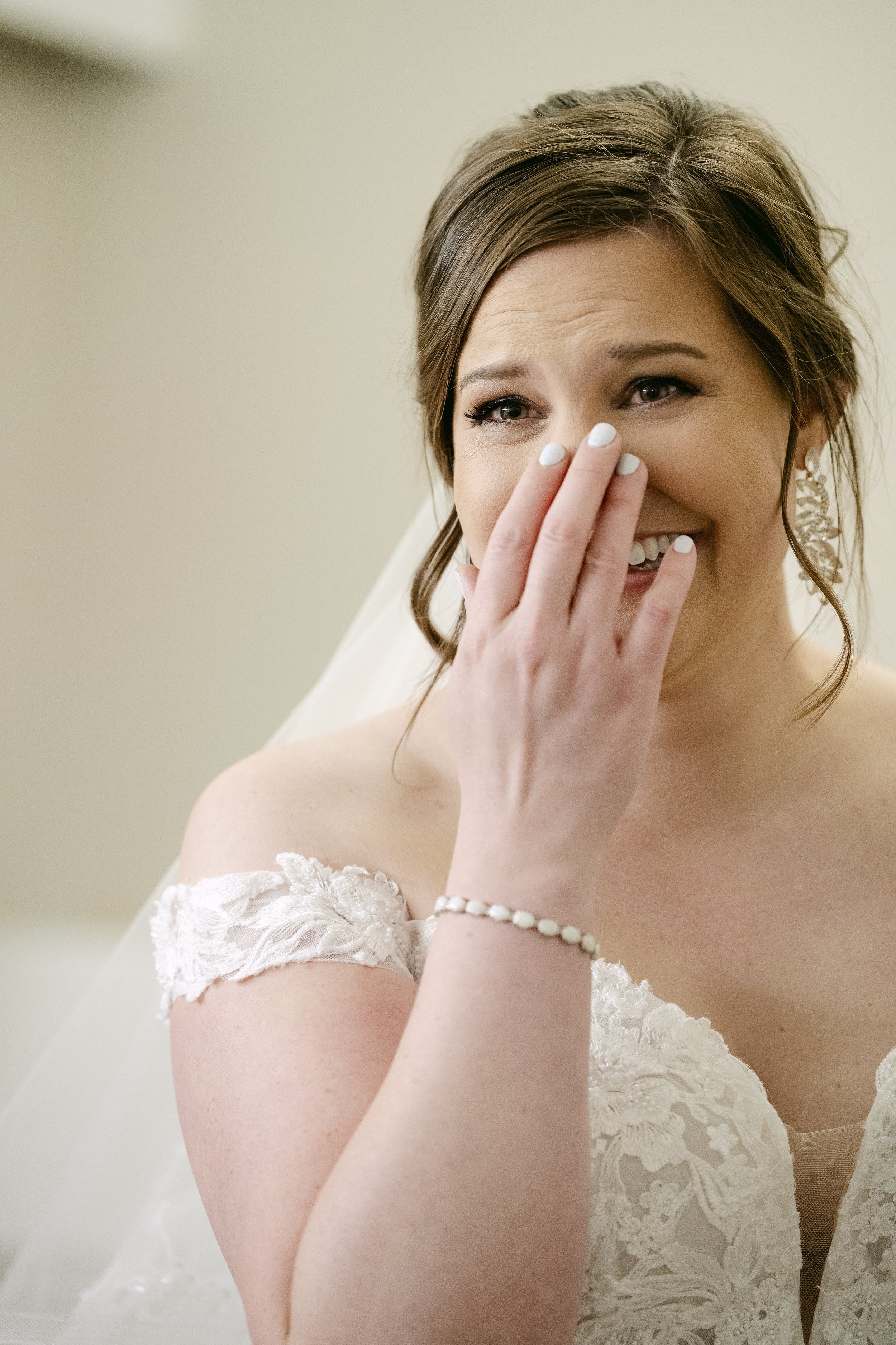 A woman in a lace wedding dress smiling with her hand covering her mouth.