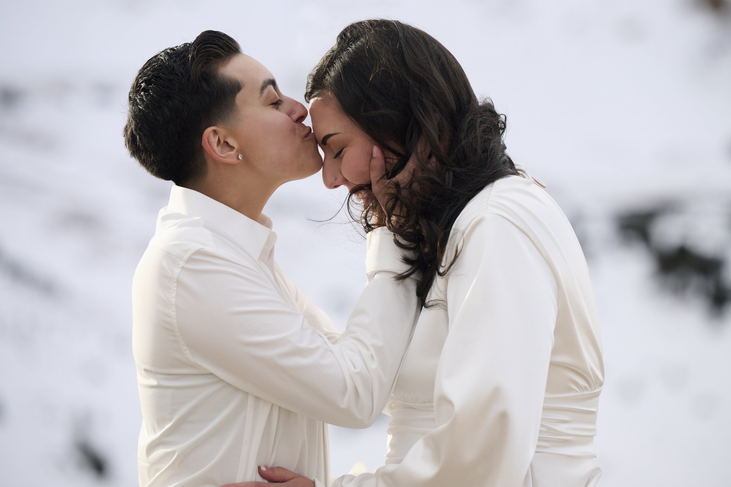 A couple sharing an intimate moment in the snow, with the woman receiving a kiss on the forehead and holding the man's face.