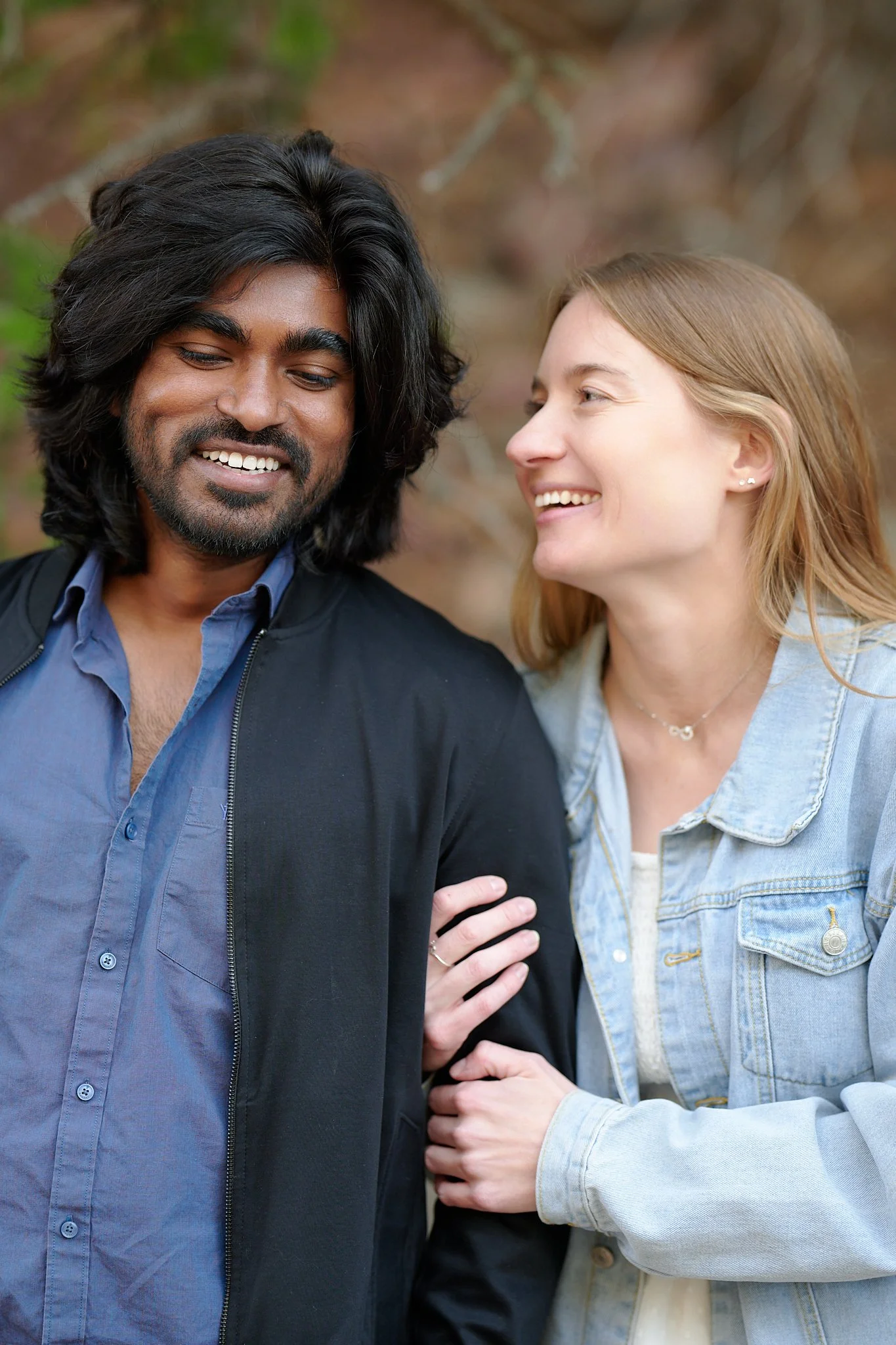 A man and woman smiling together outdoors, with the woman holding the man's arm.