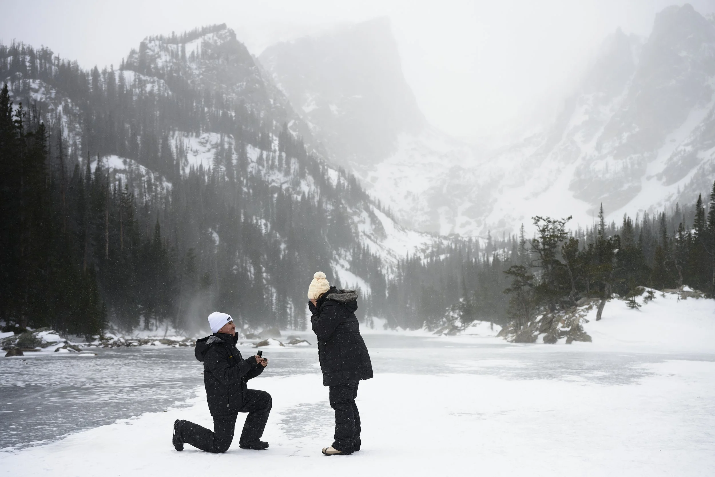 A man kneeling on one knee on snow-covered ground, proposing with a ring box to a woman who is standing with her hand on her face in a snowy mountain landscape with trees and a frozen lake.