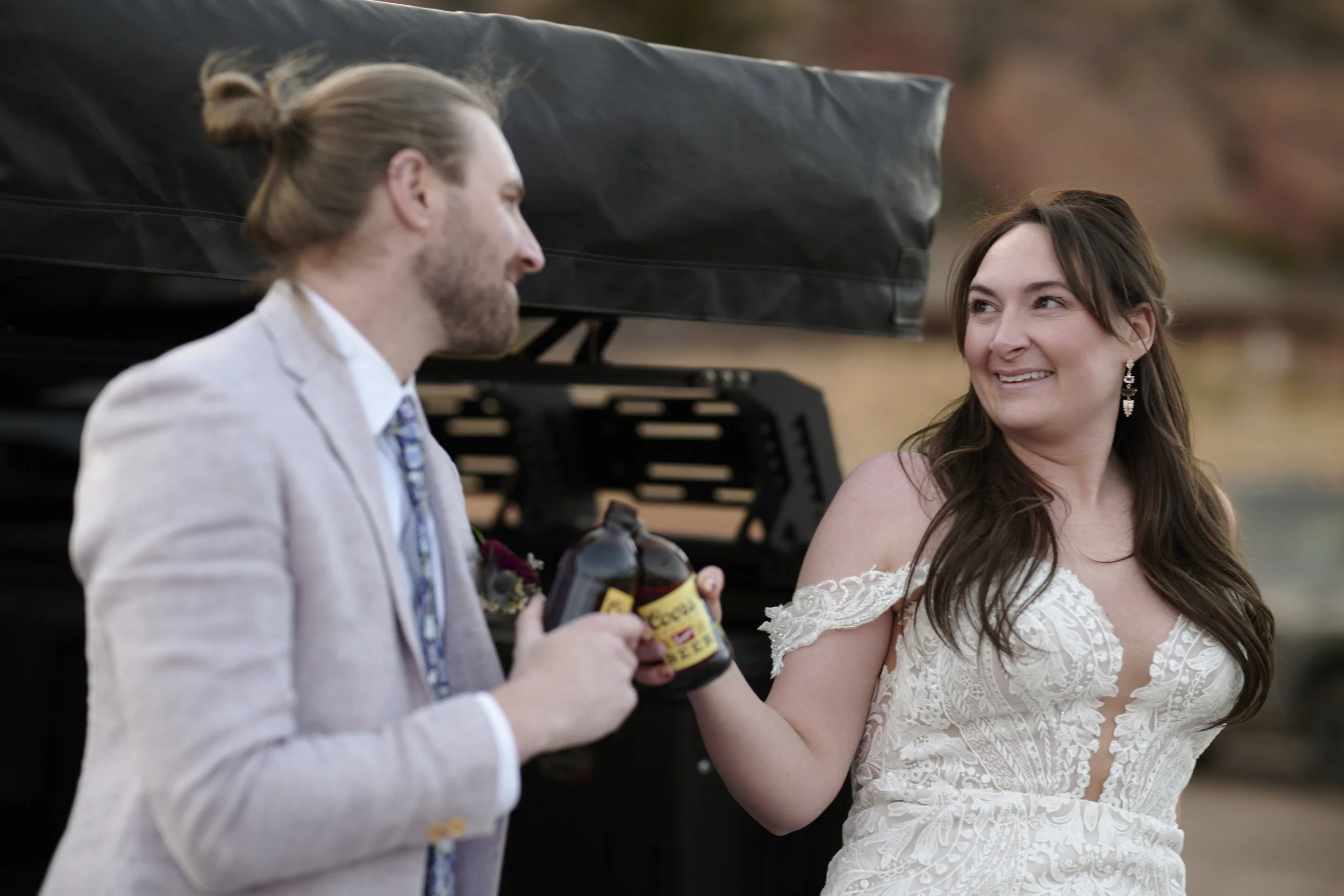 A man in a light suit holding a bottle of alcohol, smiling at a woman in a bridal dress, outdoors near a body of water.