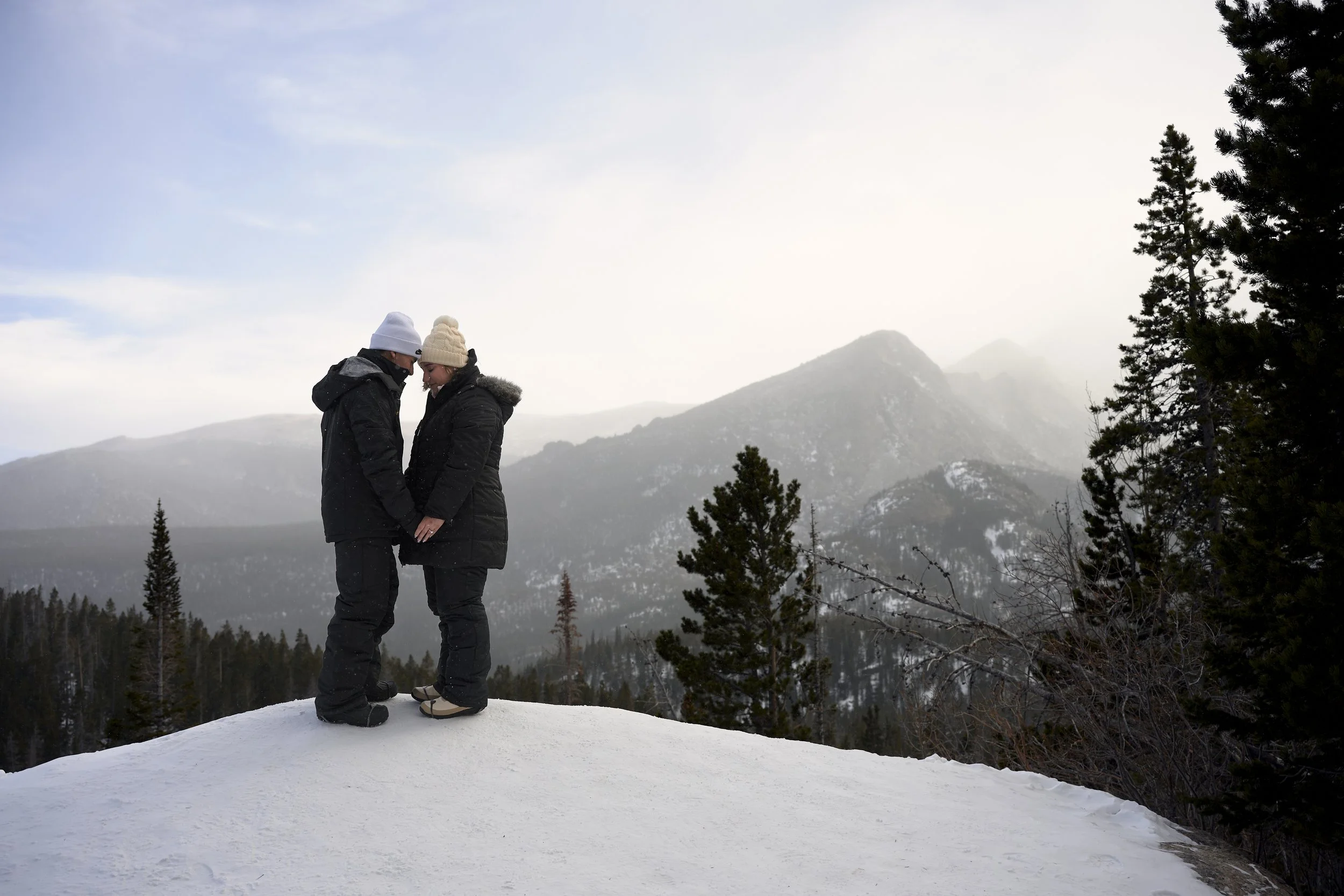 A couple poses on an overlook with a mountain backdrop while hiking to the lake for their Dream Lake engagement photos