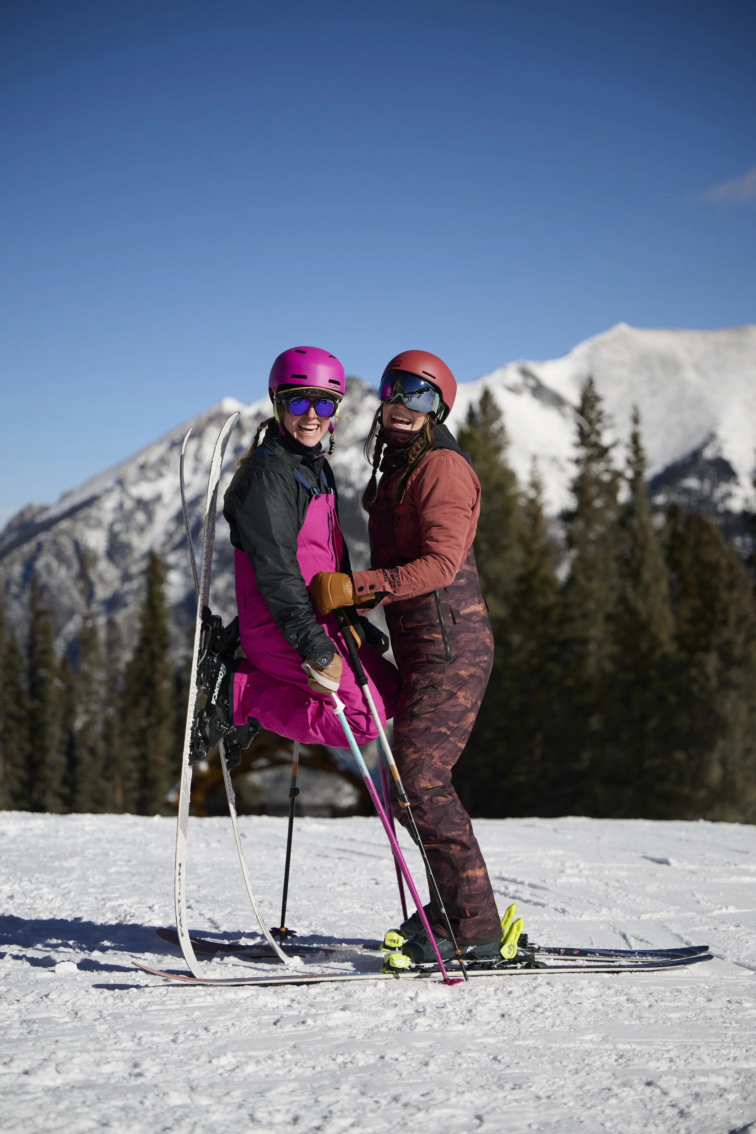 Two women in winter sports gear smiling and holding each other on skis in a snowy mountain landscape with trees and snow-covered peaks in the background.