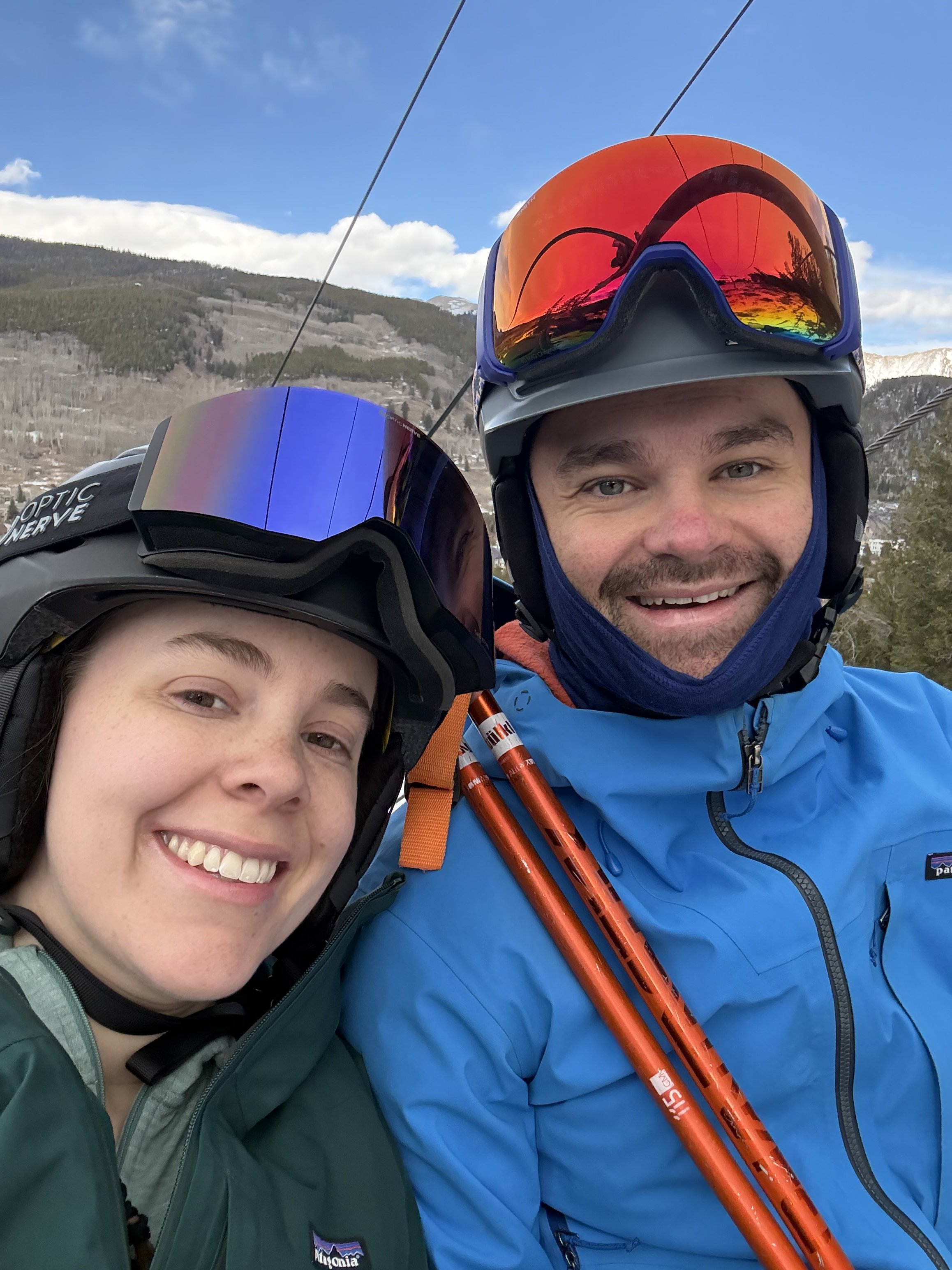 A man and a woman in ski gear taking a selfie on the ski slope with snowy mountains and a blue sky in the background.