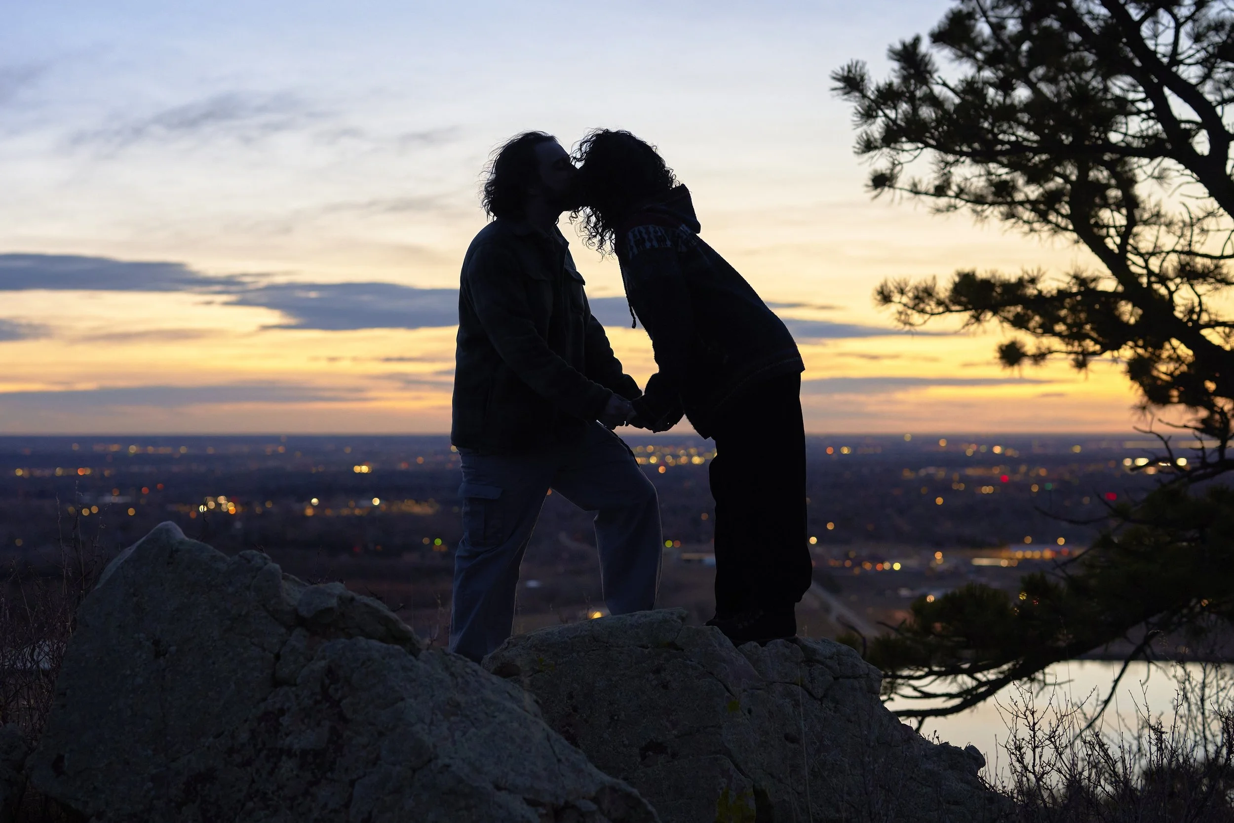 A couple sharing a kiss and holding hands on a rocky hilltop during sunset with city lights in the background.