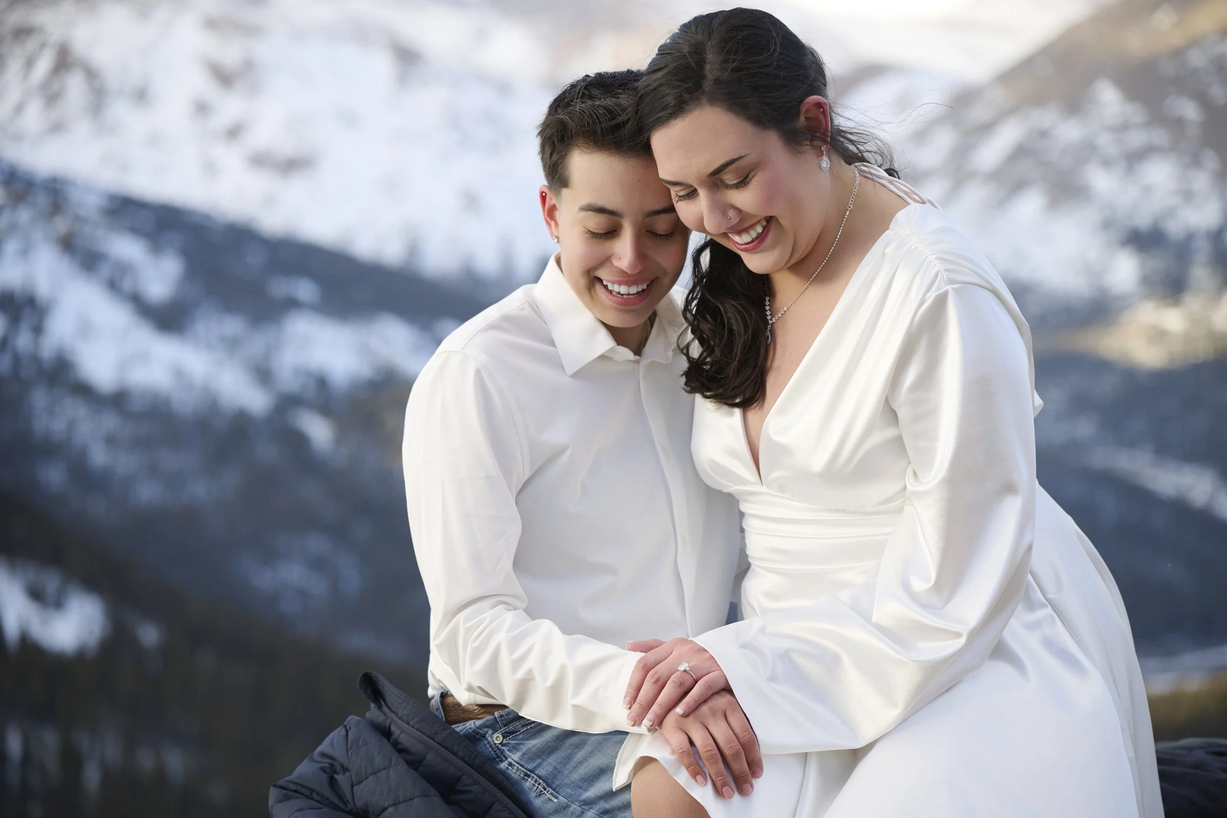 Two women, one in a white dress and one in a white shirt and jeans, sitting close together outdoors with snowy mountains in the background, smiling and holding hands.