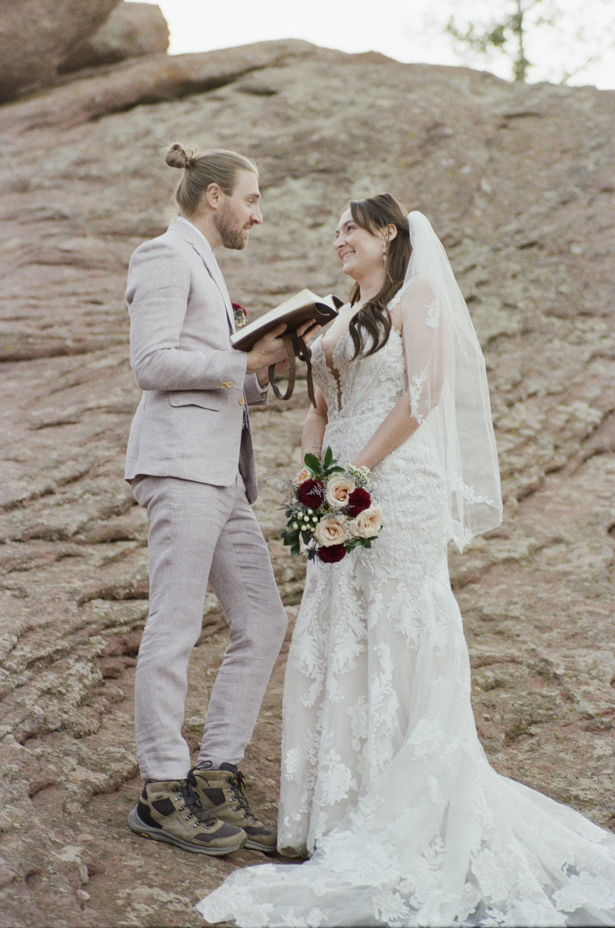 A bride and groom exchange vows outdoors on rocky terrain, with the groom holding a book and the bride holding a bouquet of flowers, both smiling at each other.