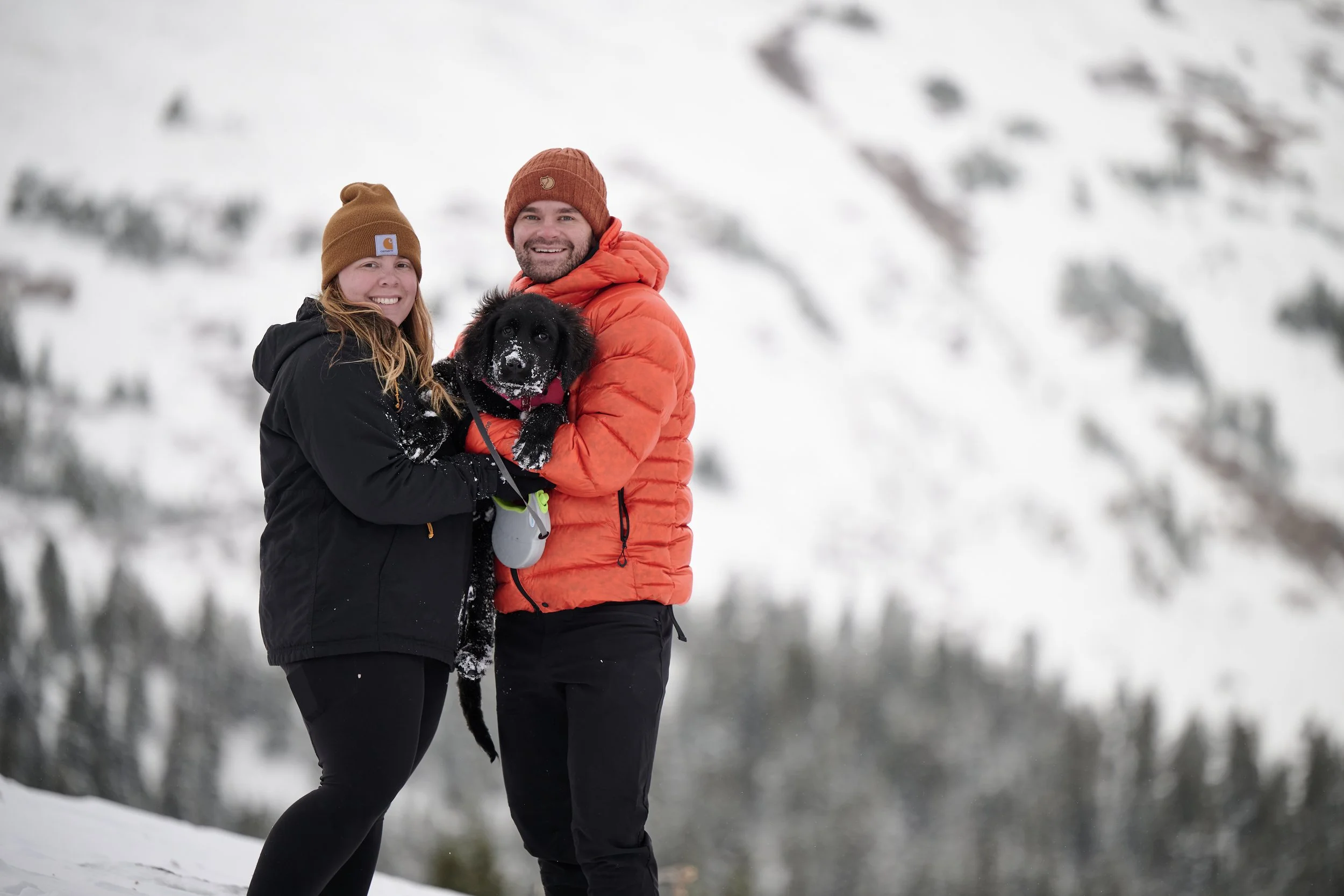 A man and woman in winter clothing standing outdoors in a snowy landscape, holding a black puppy between them, both smiling, with snow-covered trees in the background.