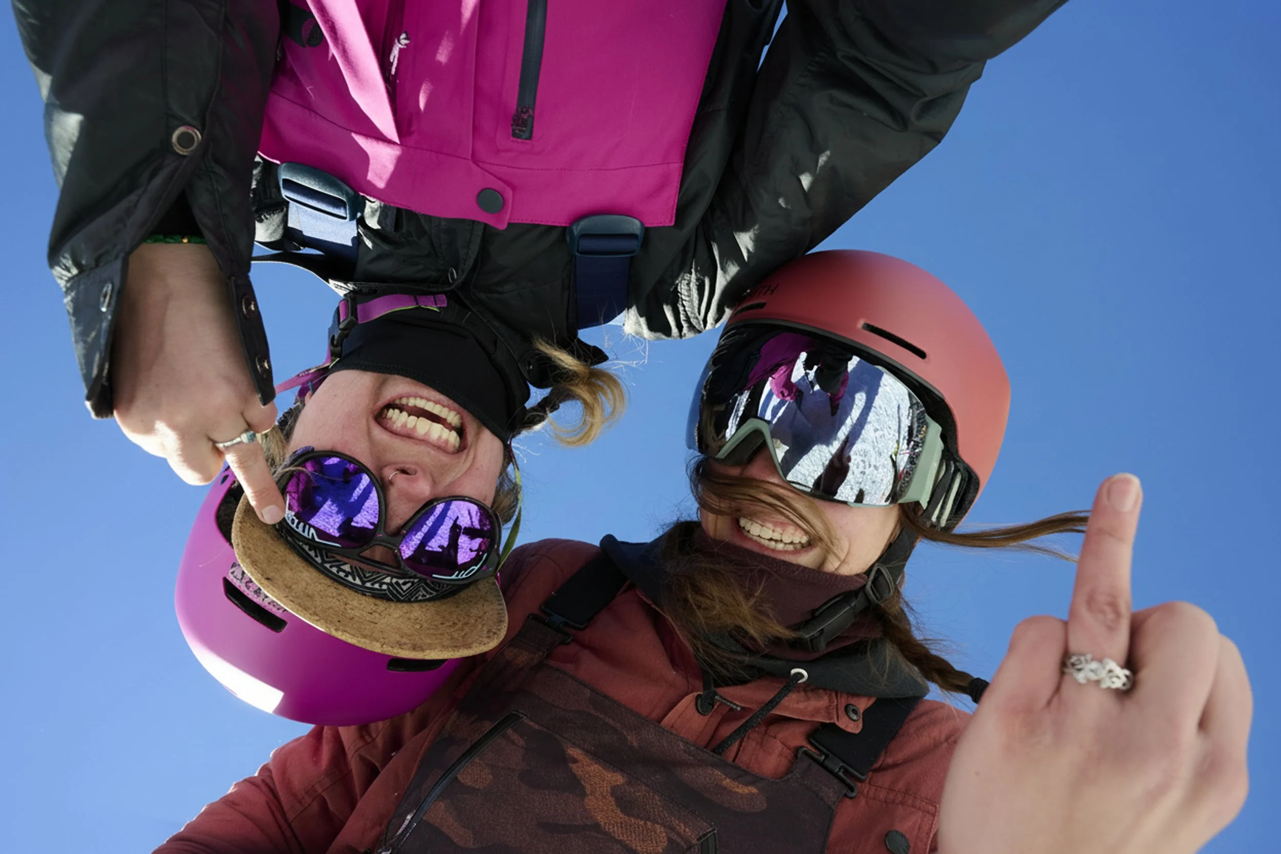 Two smiling women, one in pink snowboard gear with a pink helmet and goggles, the other in a red jacket with a red helmet and reflective goggles, posing upside down for a selfie against a clear blue sky, both showing their middle fingers.