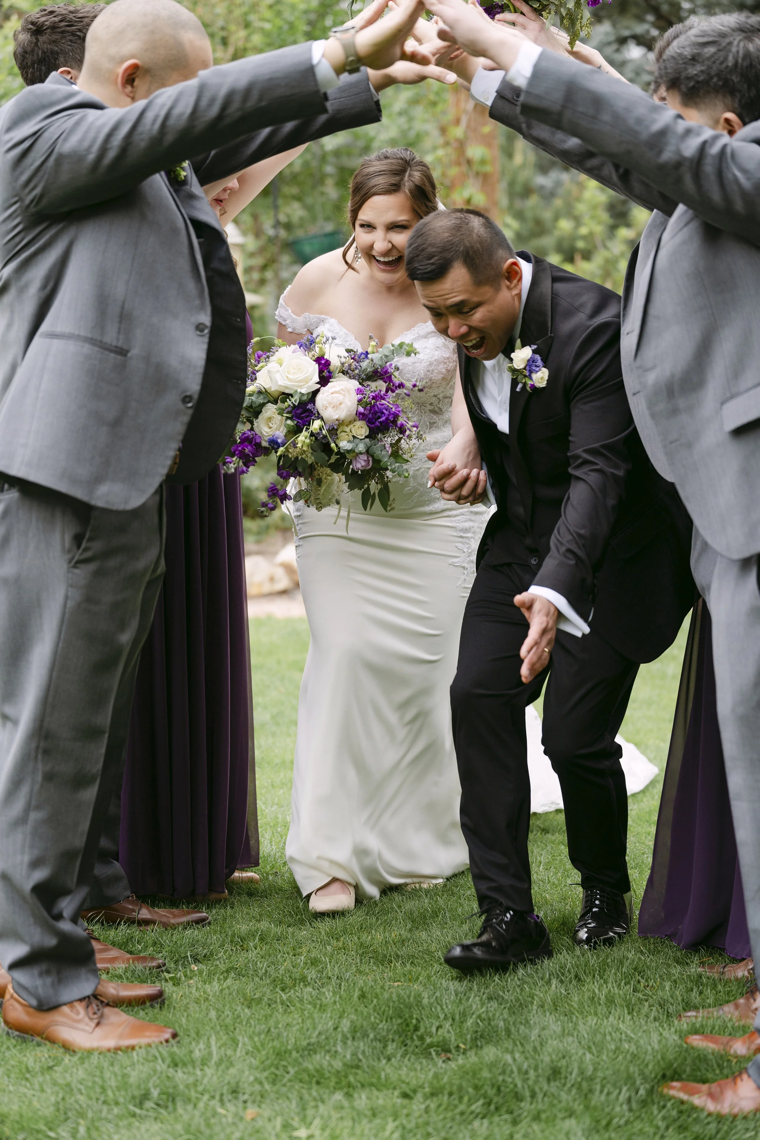 A bride and groom with friends standing in a circle outdoors, with the friends forming a tunnel with their arms, as the couple walks through holding hands. The bride is wearing a white wedding dress and holding a bouquet of flowers, while the groom i
