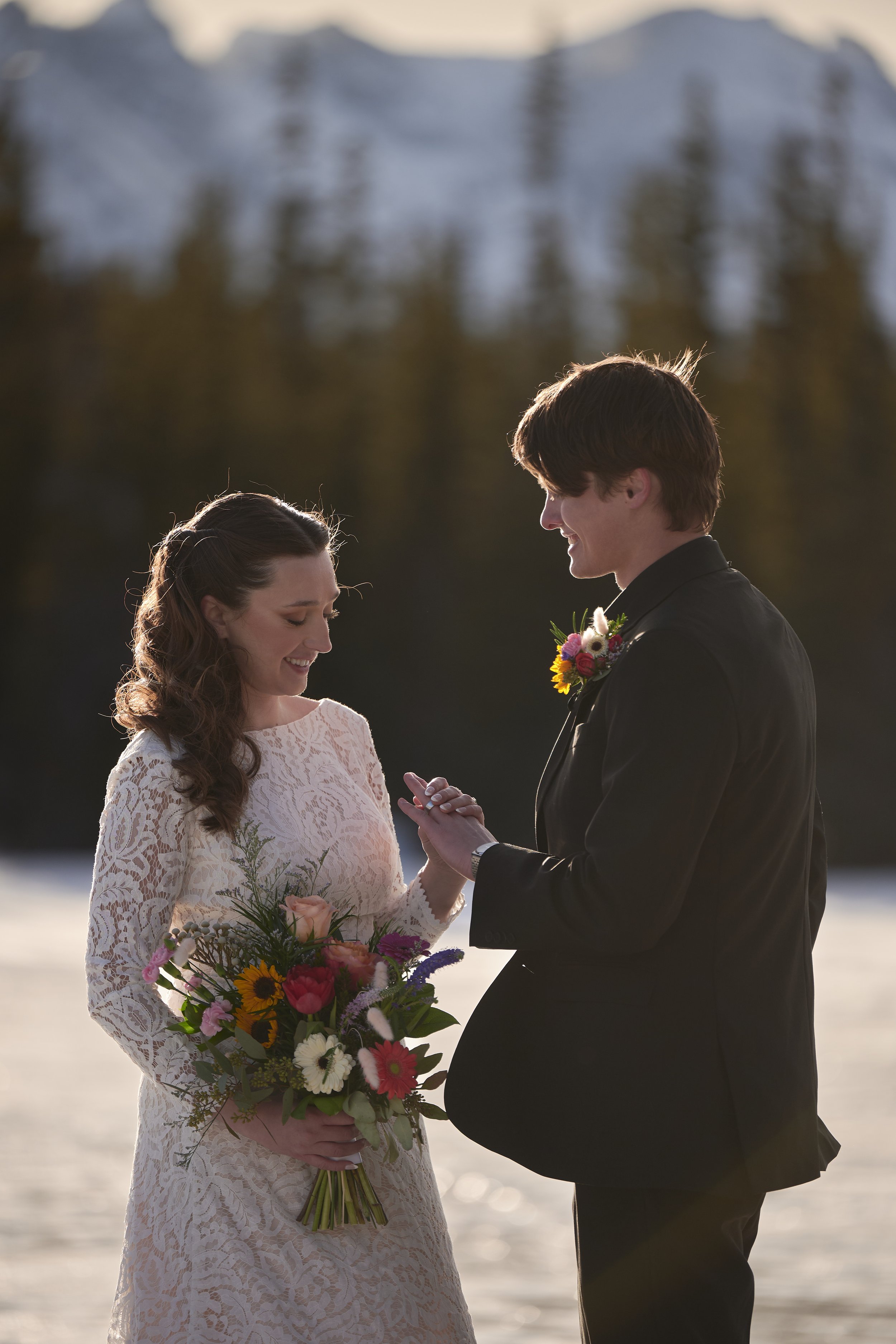 A bride and groom standing outdoors during their wedding ceremony, holding hands and smiling at each other, with the bride holding a colorful bouquet of flowers.